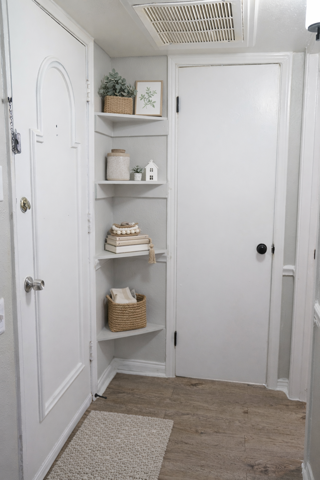 A light-filled entryway featuring a white door, a small shelf with decorative items like potted plants and a candle, and a closed door with a simple black handle. The wooden floor is partially covered by a textured rug, adding warmth to the space. The walls are painted in neutral tones.