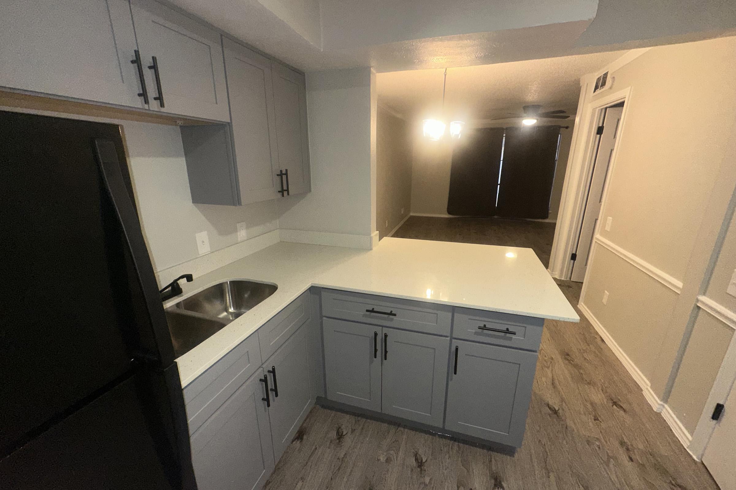 A modern kitchen featuring gray cabinets, a black refrigerator, and a white countertop. The space includes a sink and an adjoining area with a light fixture, leading to a dark carpeted room with closed curtains.