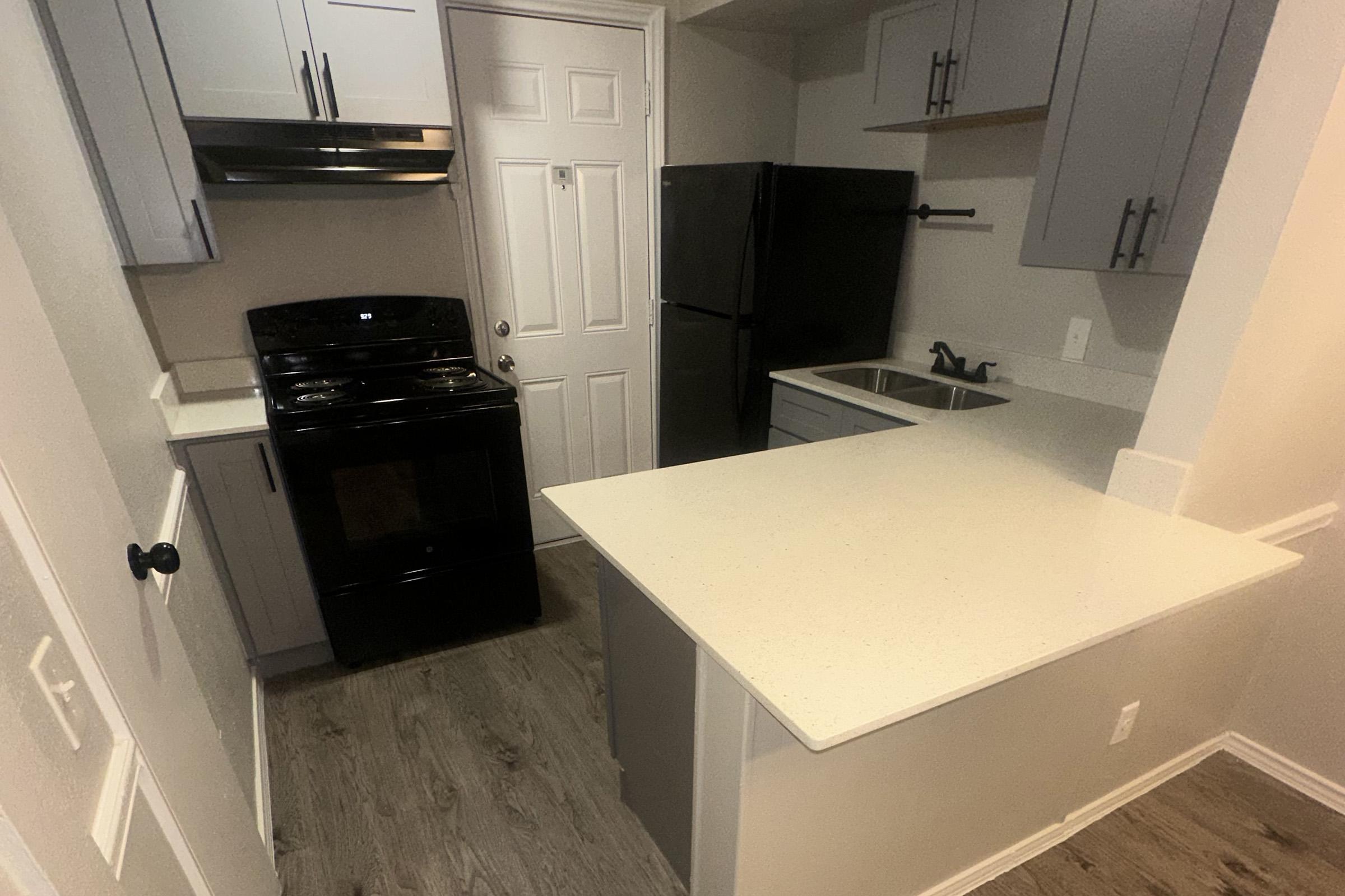 A modern kitchen featuring light gray cabinets, a black stove, and a double-door refrigerator. The countertop is white and extends into a small breakfast bar. The space is illuminated with warm lighting, and there is a door visible that leads outside. The flooring is a light wood tone, enhancing the contemporary design.