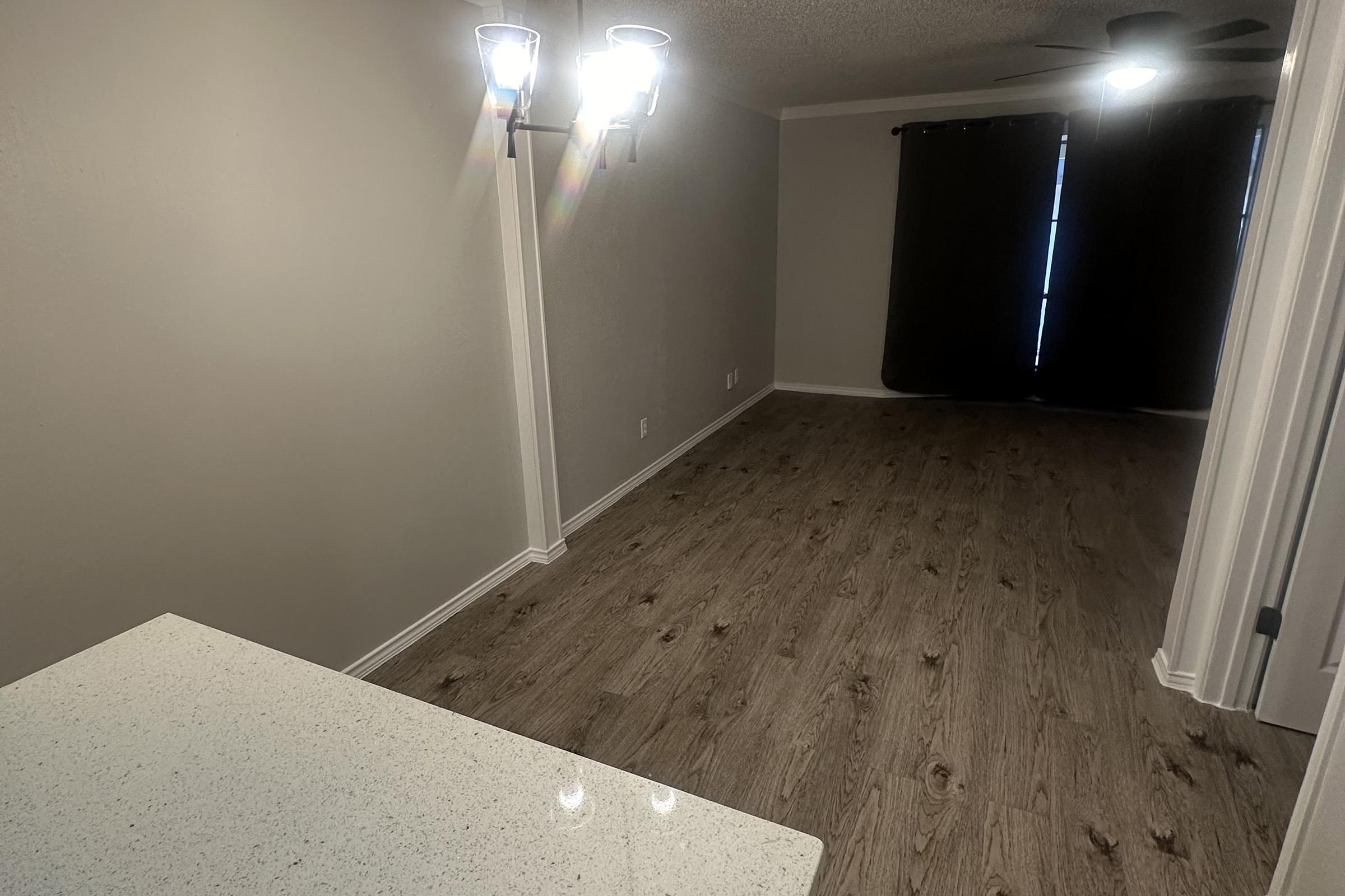 A spacious, empty room with light-colored walls and wooden flooring. There are two wall-mounted light fixtures illuminating the space. A dark curtain partially obscures a window, and a ceiling fan is visible. In the foreground, a countertop with a speckled design is present, suggesting a kitchen or living area setup.