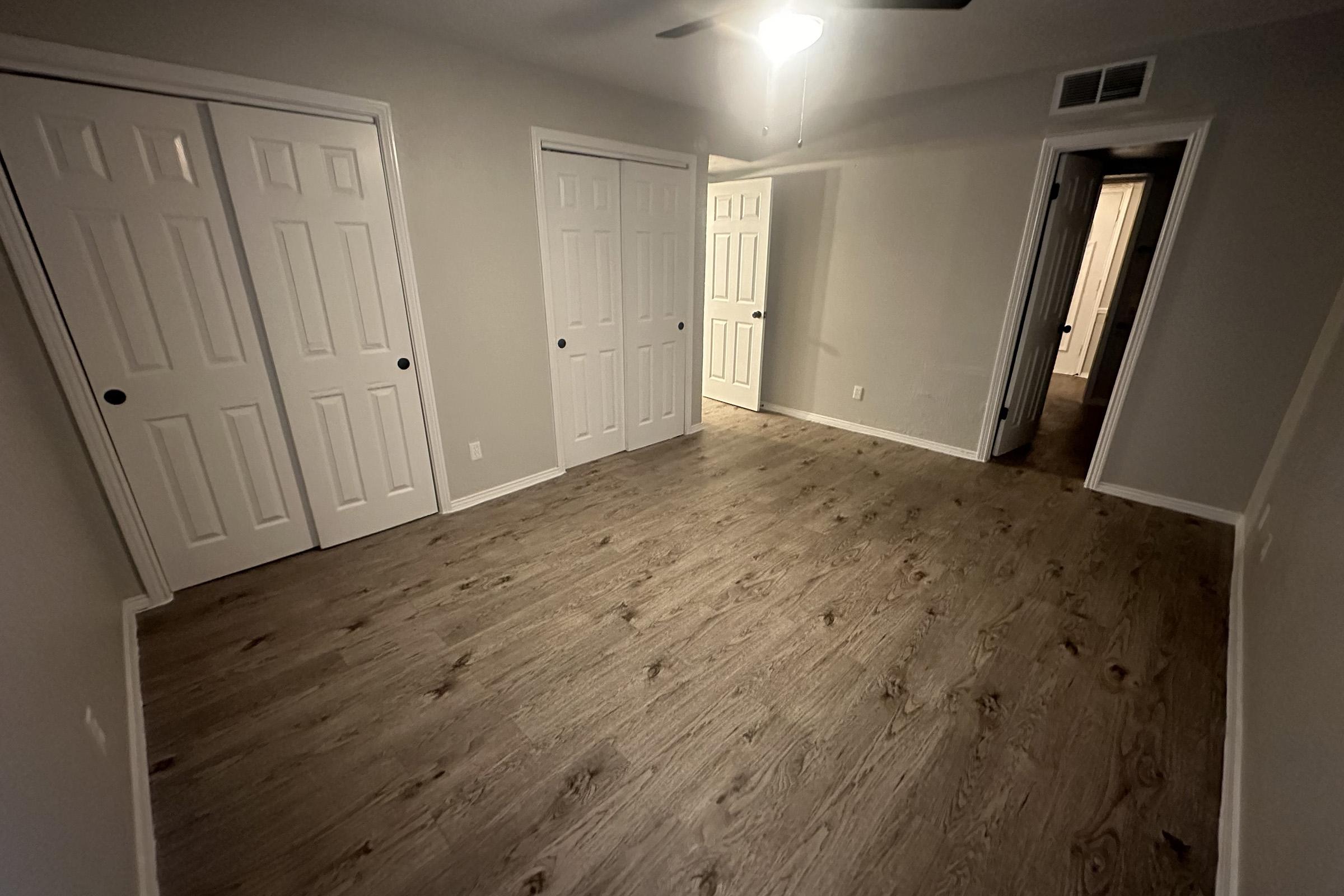 Empty room with light gray walls and a wooden floor. There are three white double doors along one wall, and another door can be seen in the background. A ceiling fan is visible, and the room is well-lit, creating a spacious and open atmosphere.