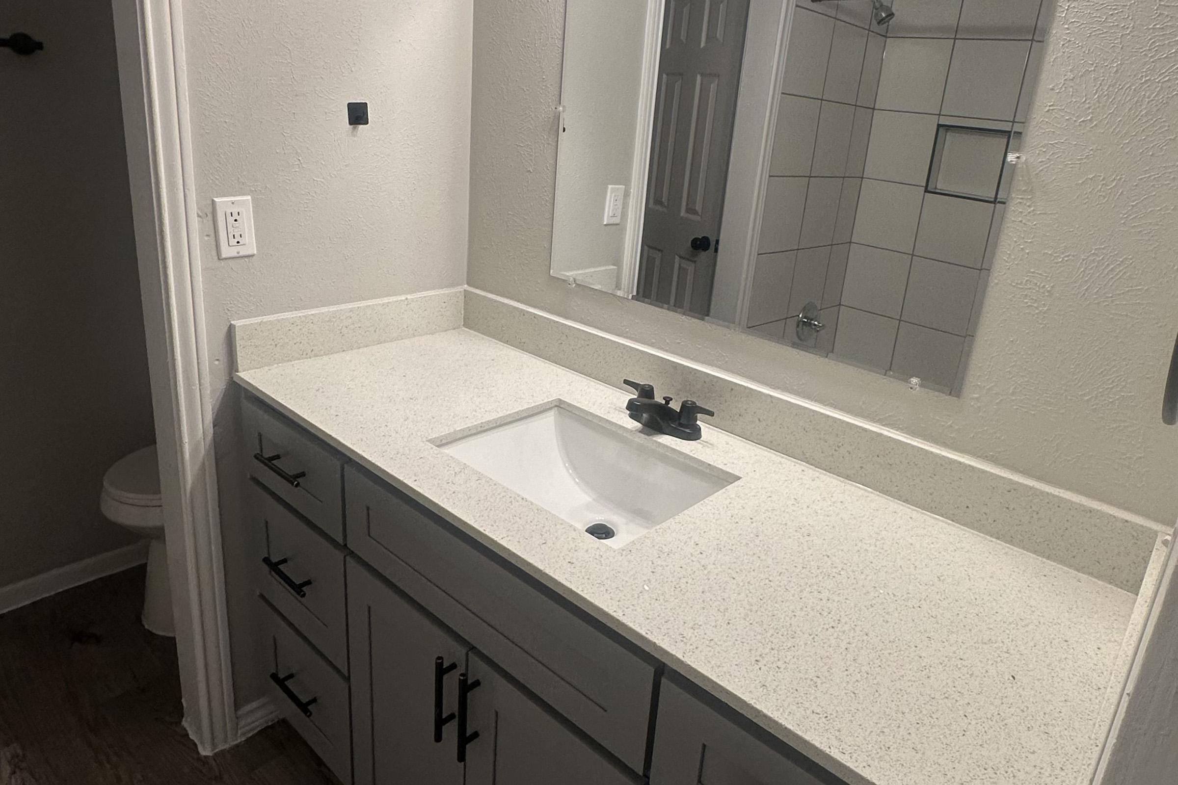 A modern bathroom vanity with a white countertop and a single sink, featuring a sleek faucet. The wall is painted a light color, and there is a large mirror above the sink. A shower area with tiled walls is visible in the background, along with a partially visible toilet in the corner.