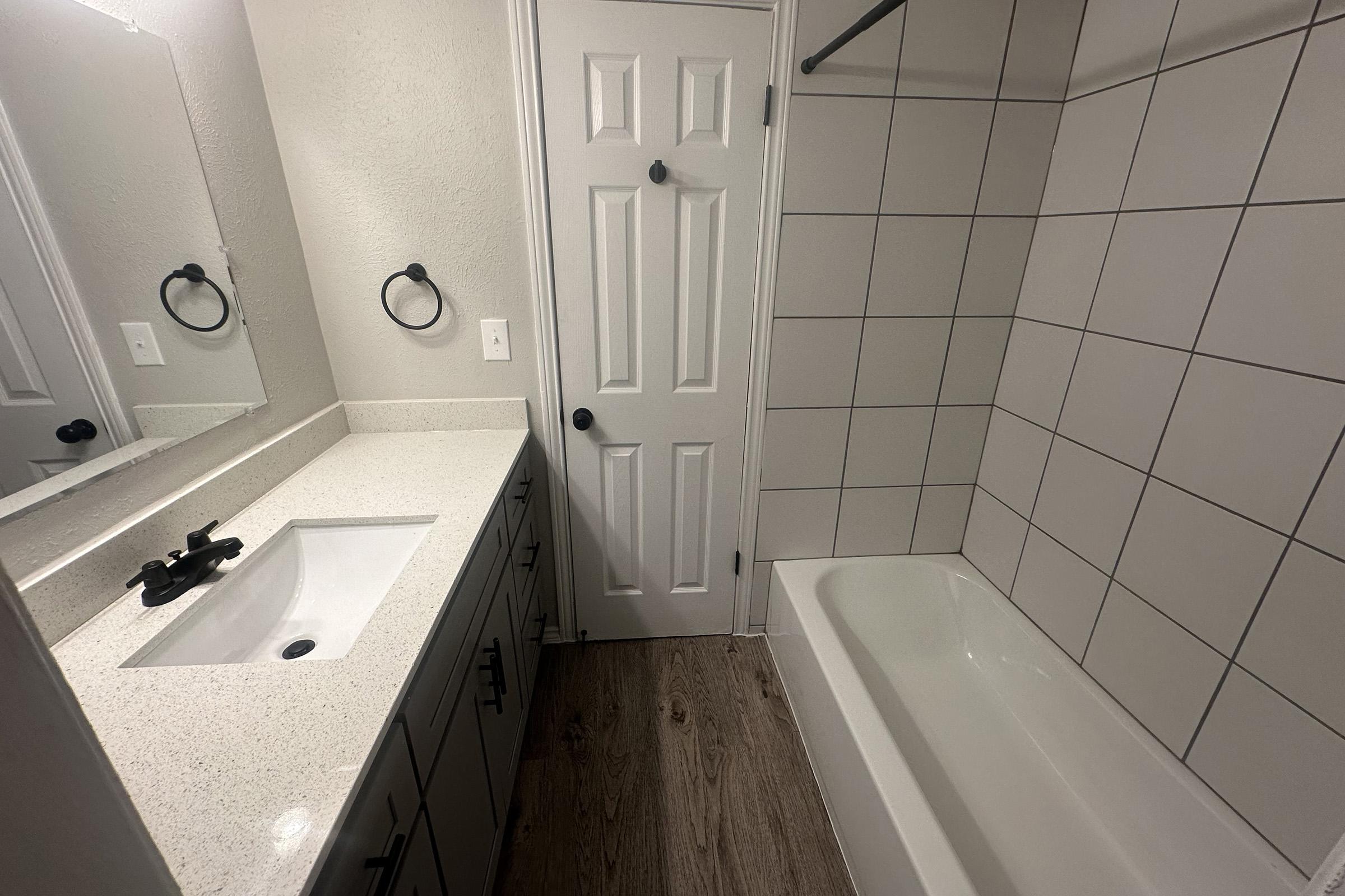 A modern bathroom featuring a white countertop with a sink, a large mirror, and dark faucet. The walls are tiled with large white tiles, and there's a bathtub along one side. A door and towel ring are visible, along with a wooden floor, creating a clean and contemporary look.