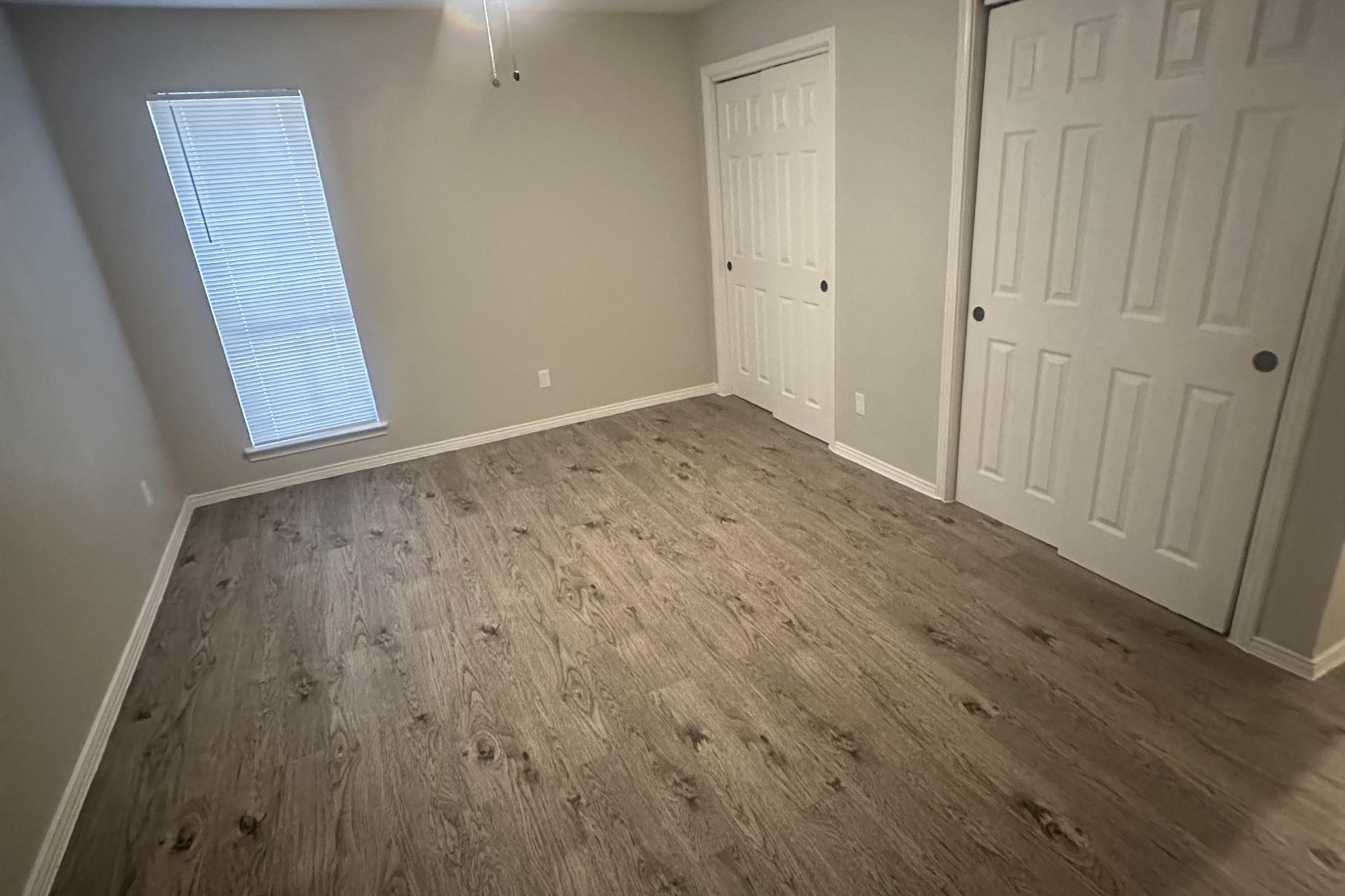 A vacant room with light gray walls and a wooden floor. There is a window with a blind partially open, allowing natural light to enter. Two sets of white closet doors are visible on one wall, creating a simple and clean aesthetic. The ceiling features a light fixture.