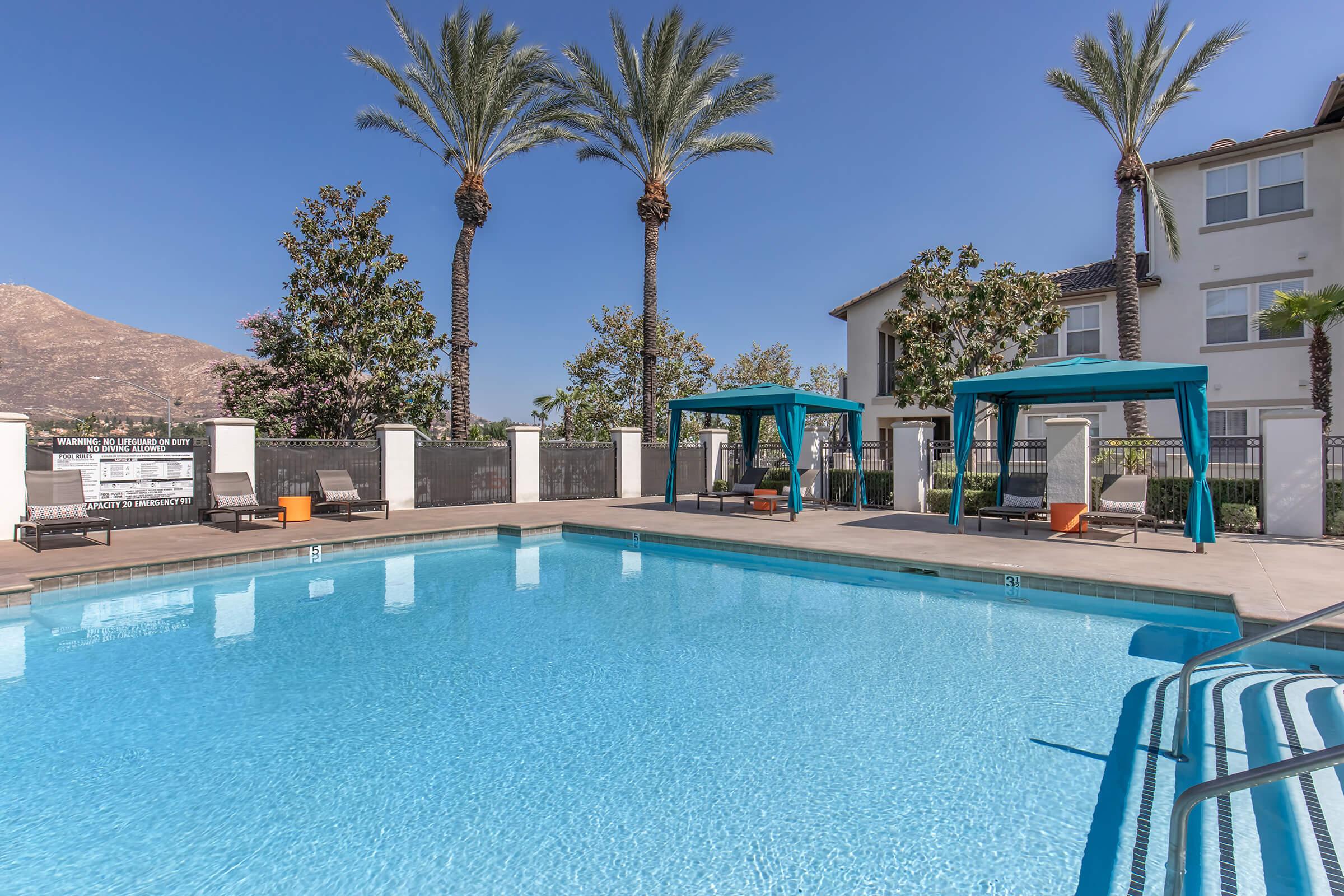 A sparkling outdoor swimming pool surrounded by lounge chairs and shaded cabanas, with palm trees and a mountain in the background. The scene is bright and inviting, perfect for relaxation and leisure.