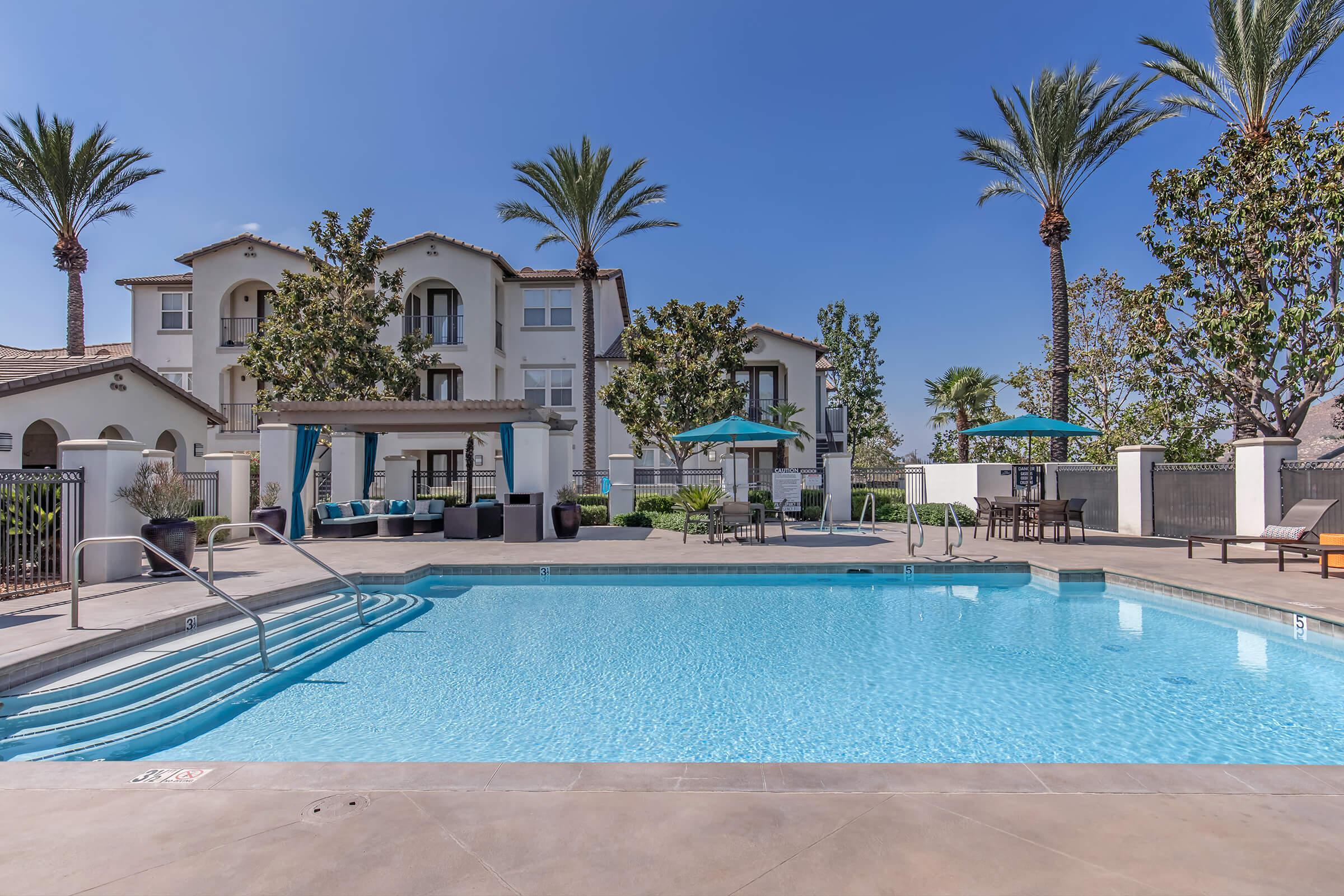 A clear blue swimming pool surrounded by palm trees and landscaped greenery. In the background, there are residential buildings with balconies. Umbrellas and lounge chairs are placed around the pool area, creating a relaxing outdoor space under a bright blue sky.