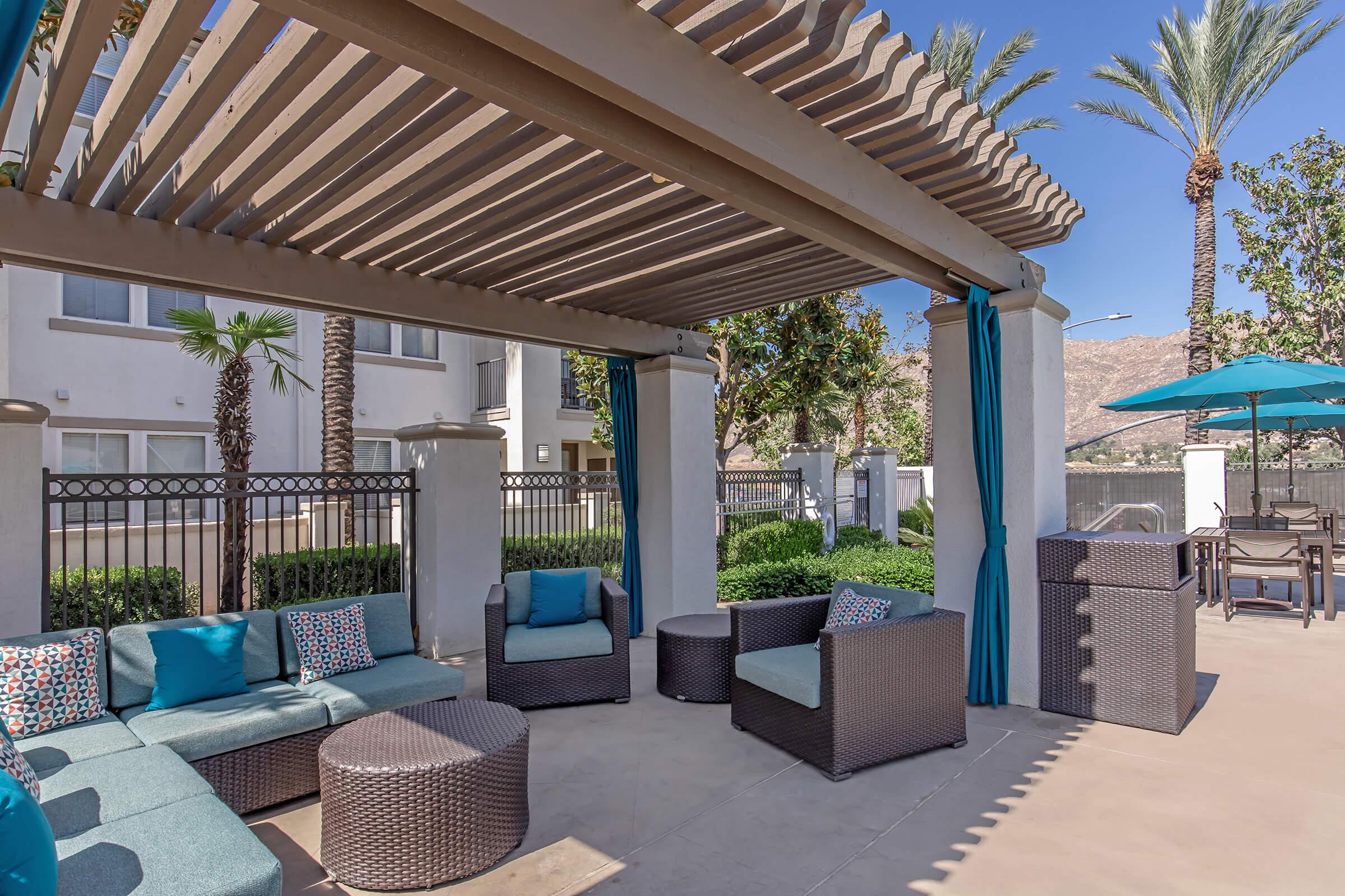Outdoor lounge area featuring a pergola with teal drapes, comfortable seating, and decorative cushions. Surrounded by palm trees and well-manicured greenery, the space is designed for relaxation, complemented by a large umbrella over a nearby table. Bright sunshine illuminates the scene.