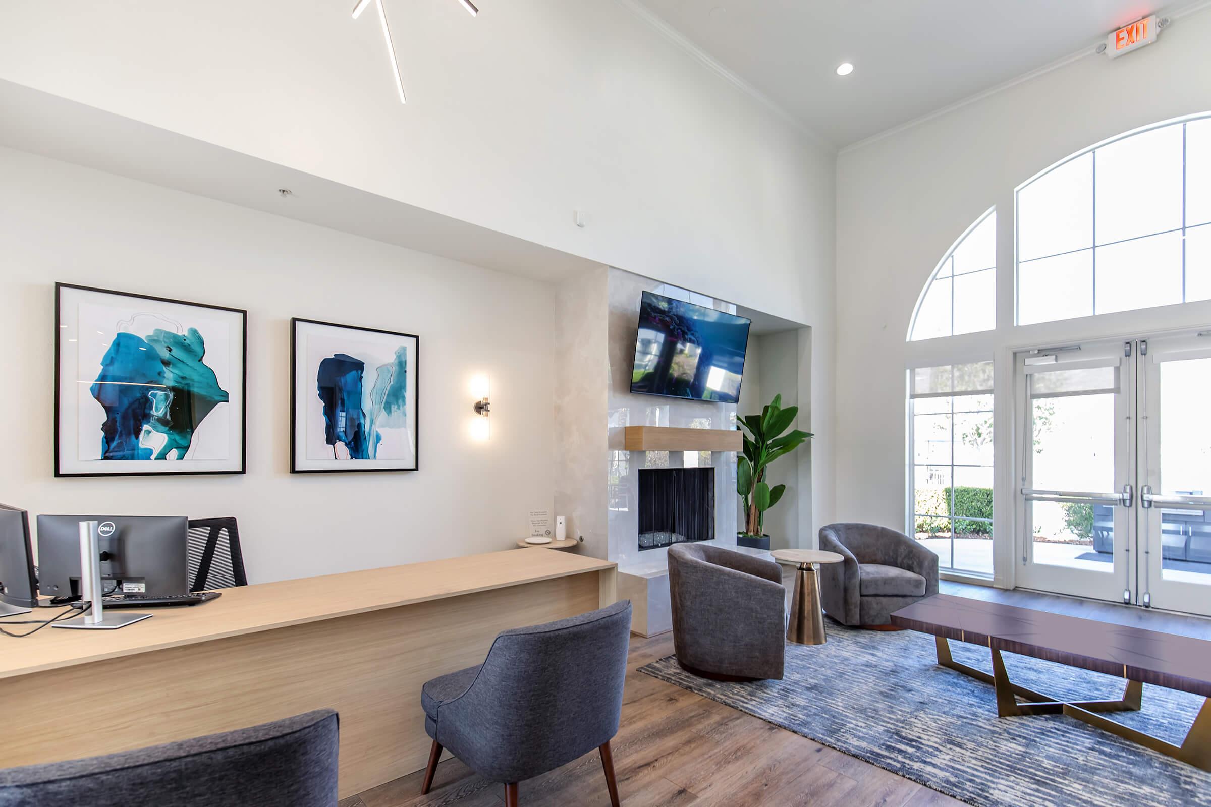 A modern reception area featuring a sleek wooden desk with a computer, two gray chairs, and a wooden coffee table. The walls are adorned with abstract art pieces in blue tones, and a large window allows natural light to brighten the space. A fireplace is visible, enhancing the cozy atmosphere.