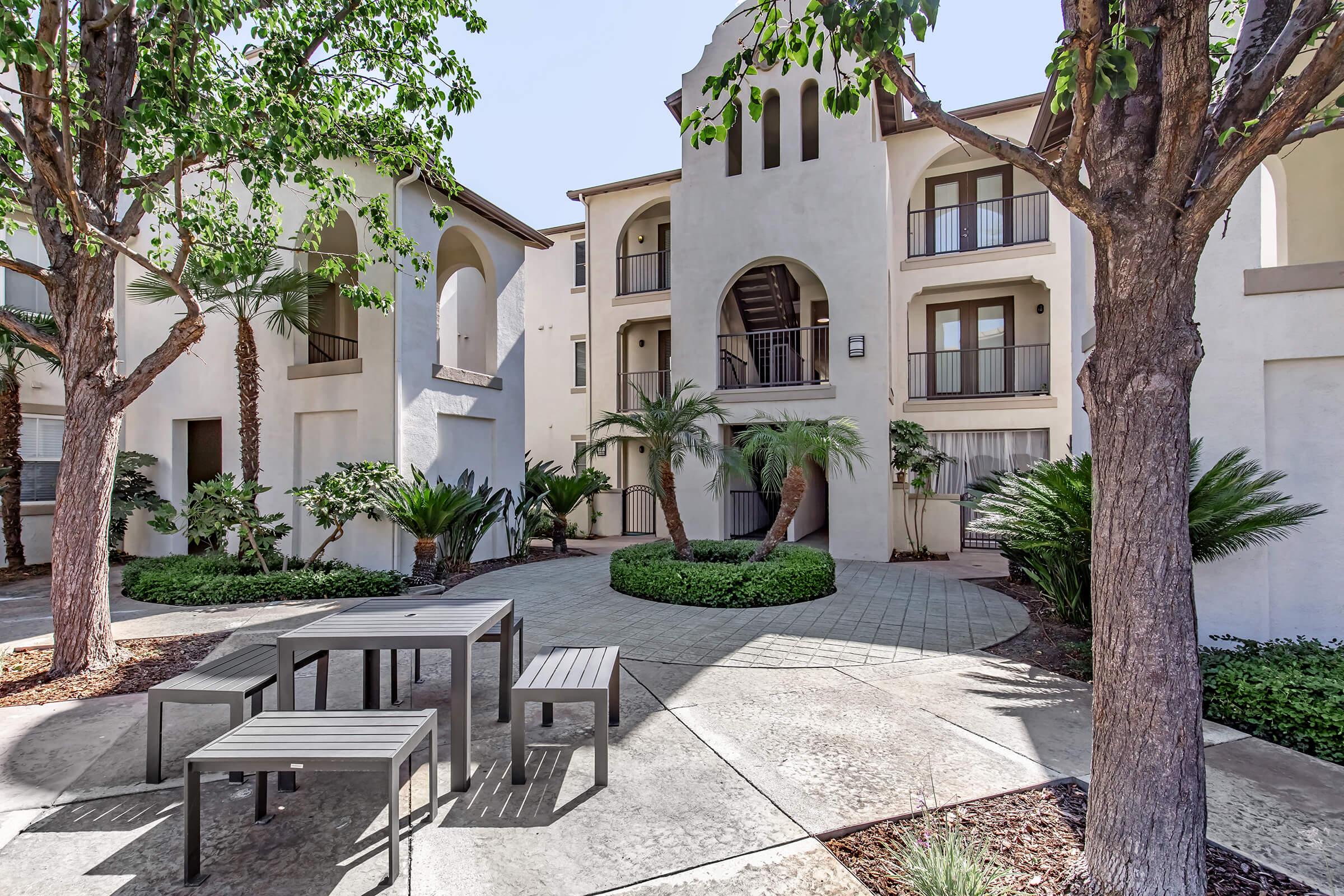 A serene courtyard featuring a patterned stone pathway surrounded by lush greenery, including palm trees and shrubs. In the center, there is a modern outdoor seating area with a sleek table and benches, against a backdrop of spacious white apartment buildings.