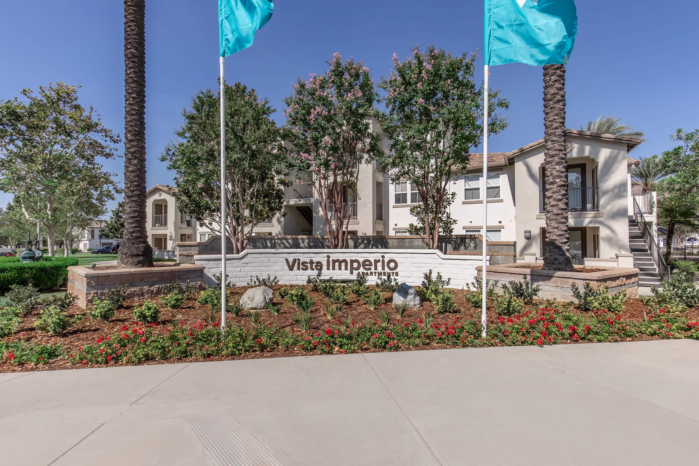 A well-maintained apartment complex entrance featuring a sign that reads "Vista Imperio." The area is landscaped with flowers and shrubs, and tall palm trees flank the entrance. Two turquoise flags are displayed on poles in front of the building, contributing to a welcoming atmosphere.