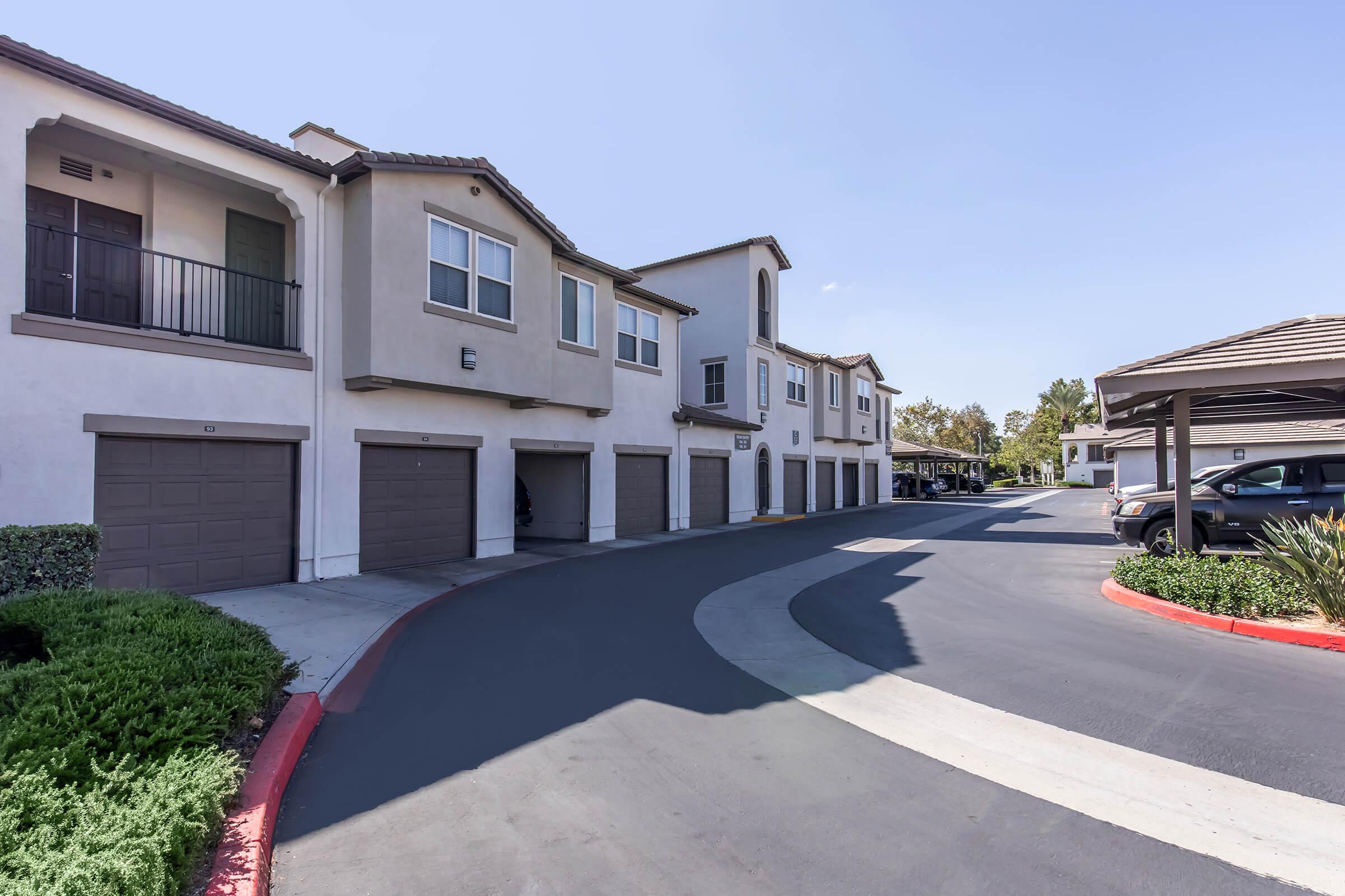 A view of a residential complex featuring two-story townhouses with attached garages. The buildings have light-colored exteriors and balconies, surrounded by well-maintained landscaping. A curved driveway leads to parking areas, with a clear blue sky overhead. The scene is bright and inviting.