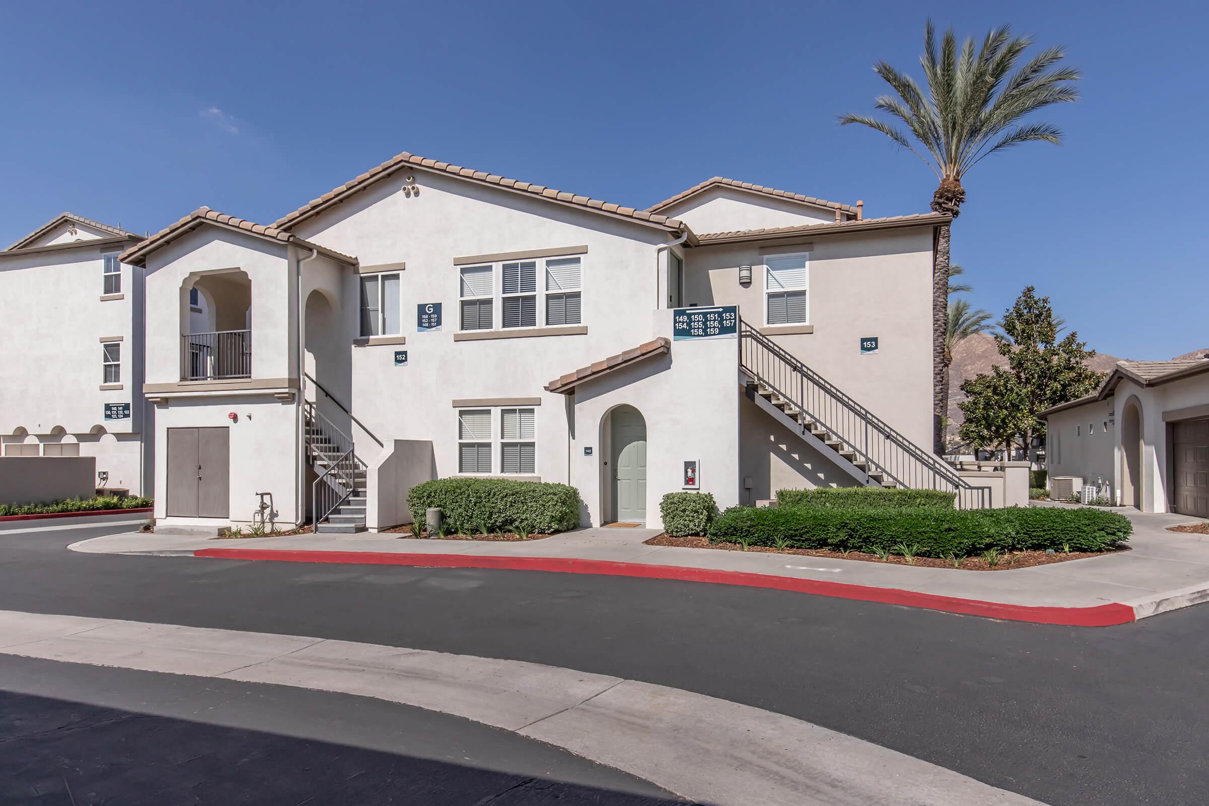 Two-story residential building with a light-colored exterior, featuring a balcony, stairs, and several windows. Palm trees are visible in the background, and the surrounding area includes a well-maintained landscape with shrubs and a circular driveway. Bright blue sky overhead.