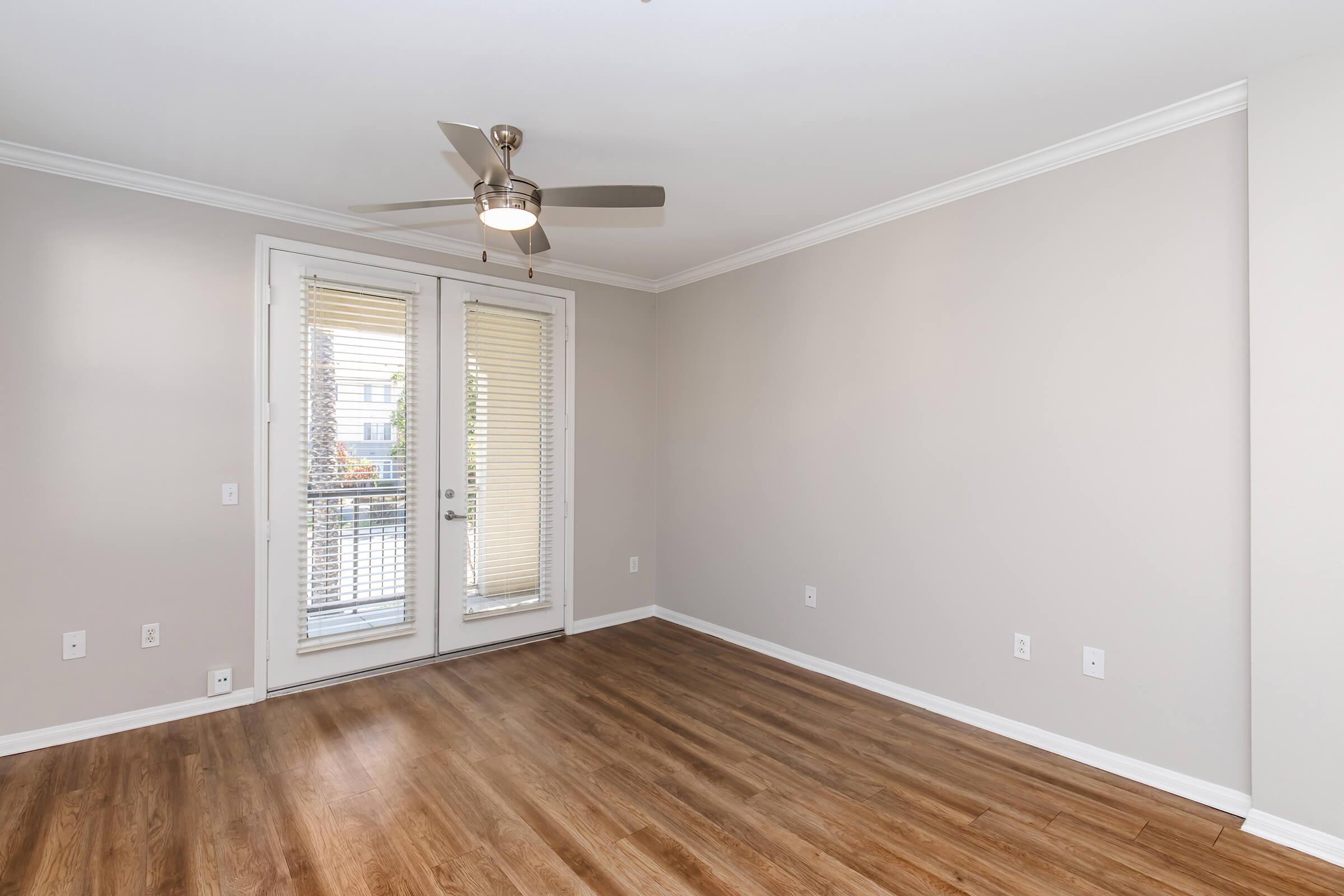 Bright and airy room with light gray walls and hardwood flooring. It features a ceiling fan and large windows with blinds, allowing natural light to flow in. Double French doors lead to a balcony, enhancing the space's openness and inviting atmosphere.