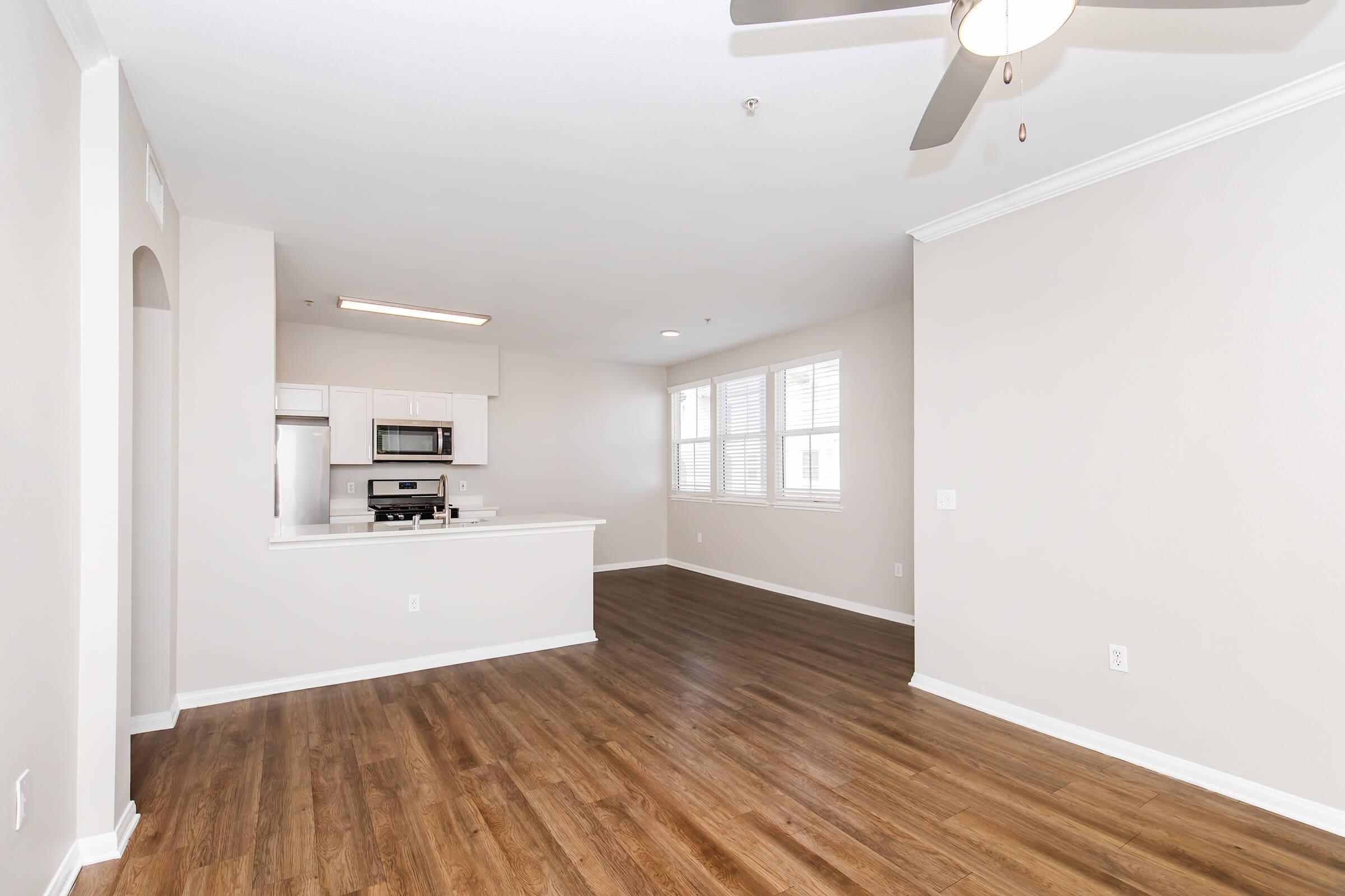 Spacious, empty living area with light-colored walls and wood-style flooring. A ceiling fan hangs above, and there are large windows allowing natural light. The adjacent kitchen features modern appliances and a breakfast bar, creating an open-concept layout.