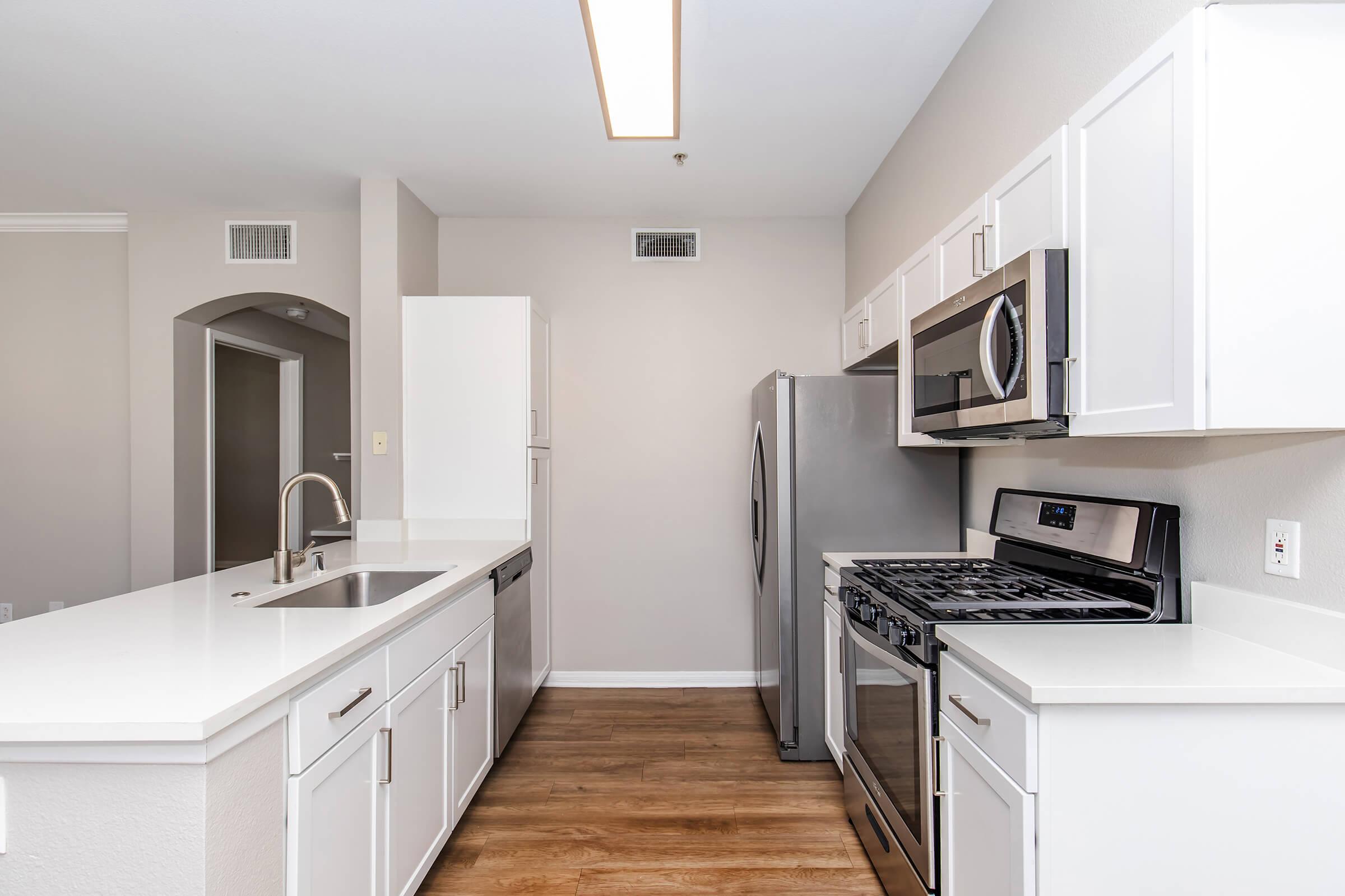 Modern kitchen featuring white cabinetry, stainless steel appliances including a gas stove and refrigerator, a double sink, and a light-colored countertop. The space has a warm wood floor and is well-lit with overhead lighting, creating a bright and inviting atmosphere.