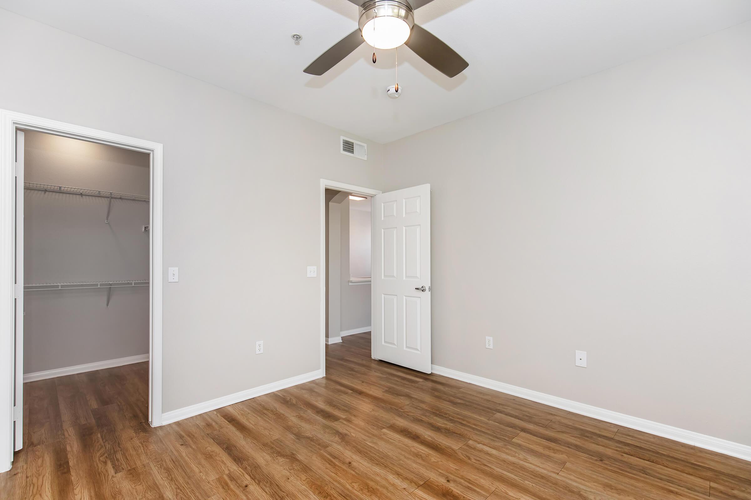A spacious room featuring hardwood floors, a ceiling fan, a door leading to a closet on the left, and another door on the right. The walls are painted in a light color, and there is a window visible in the background, allowing natural light into the space.