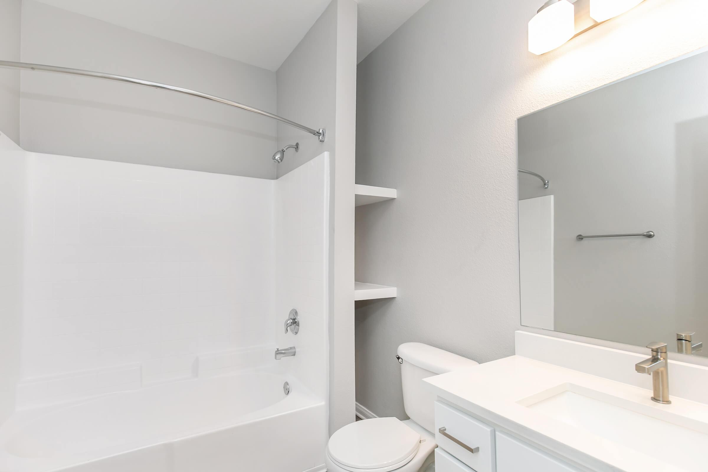 A modern bathroom featuring a white tub with a curved shower rod, a clean toilet, and a sleek vanity with a rectangular mirror. The walls are painted in a light gray color, creating a calm and spacious atmosphere. A shelf is visible next to the tub for added storage.