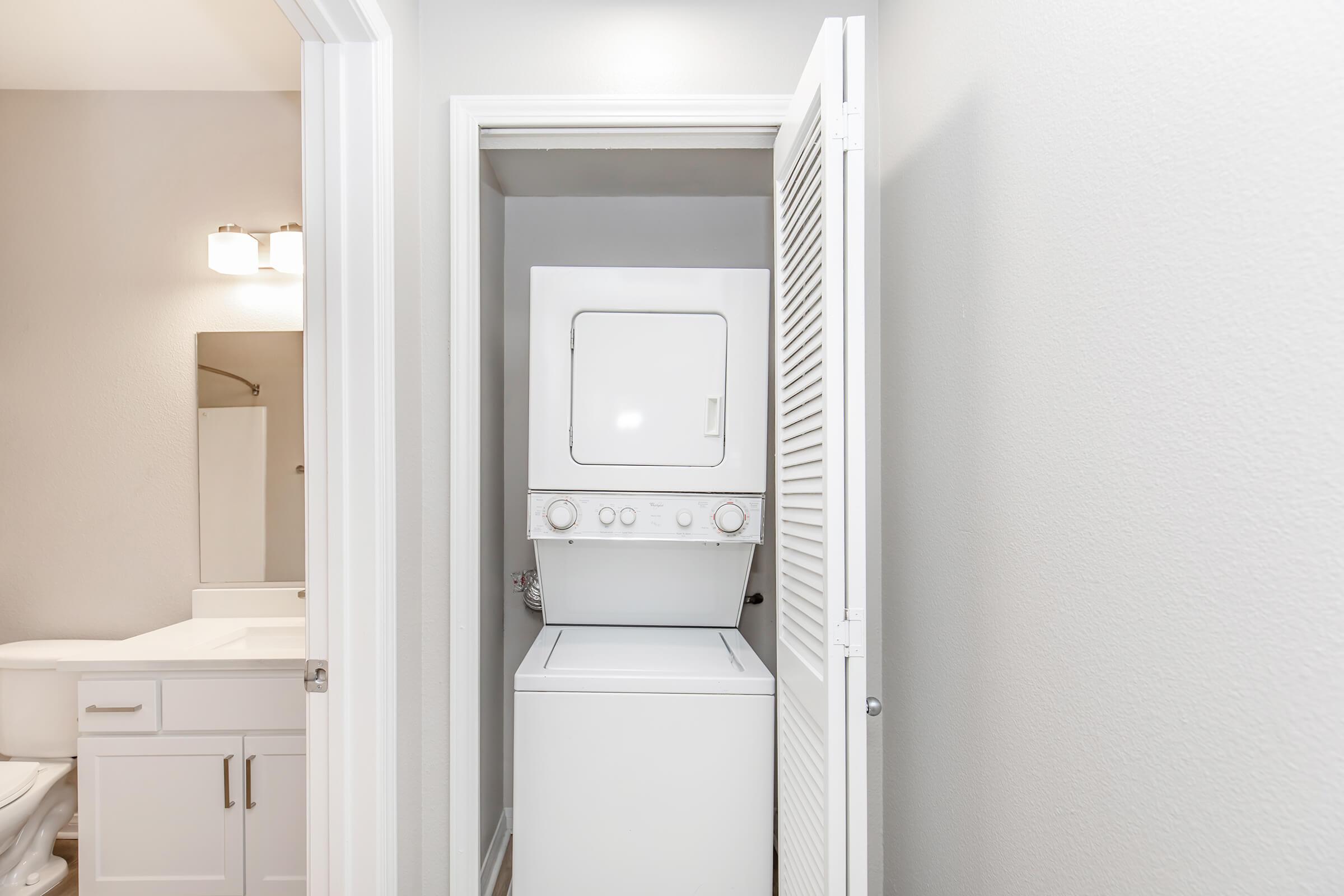 A laundry nook featuring a stacked washer and dryer within a small closet. The walls are painted light gray, and there is a bathroom visible on the left, including a white sink and mirror, with modern lighting fixtures. The overall space appears clean and well-organized.