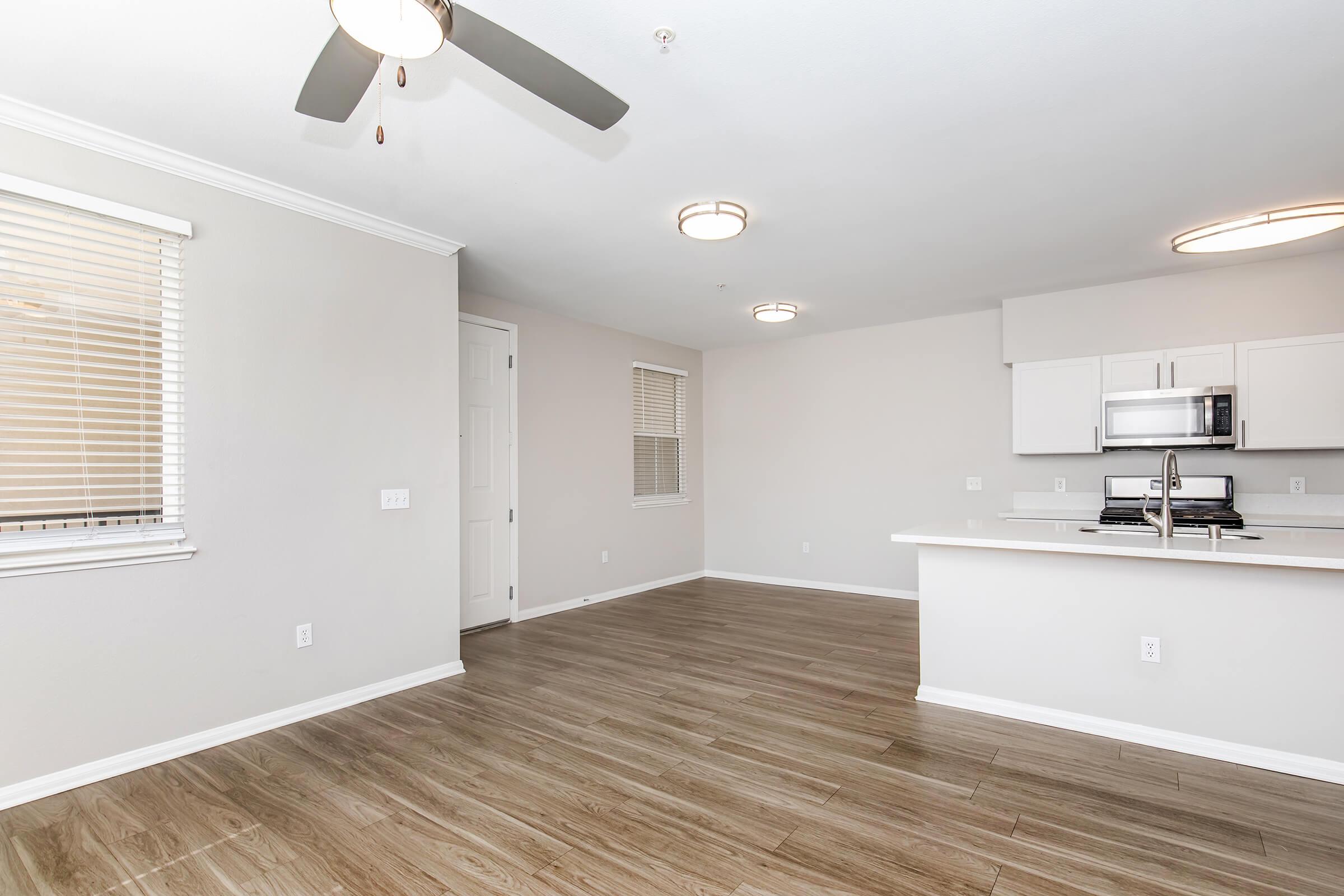 Bright and spacious living area featuring light wood flooring, neutral walls, and modern fixtures. The space includes a ceiling fan, two overhead lights, and windows with blinds. An open kitchen area with white cabinets and stainless steel appliances is visible in the background.