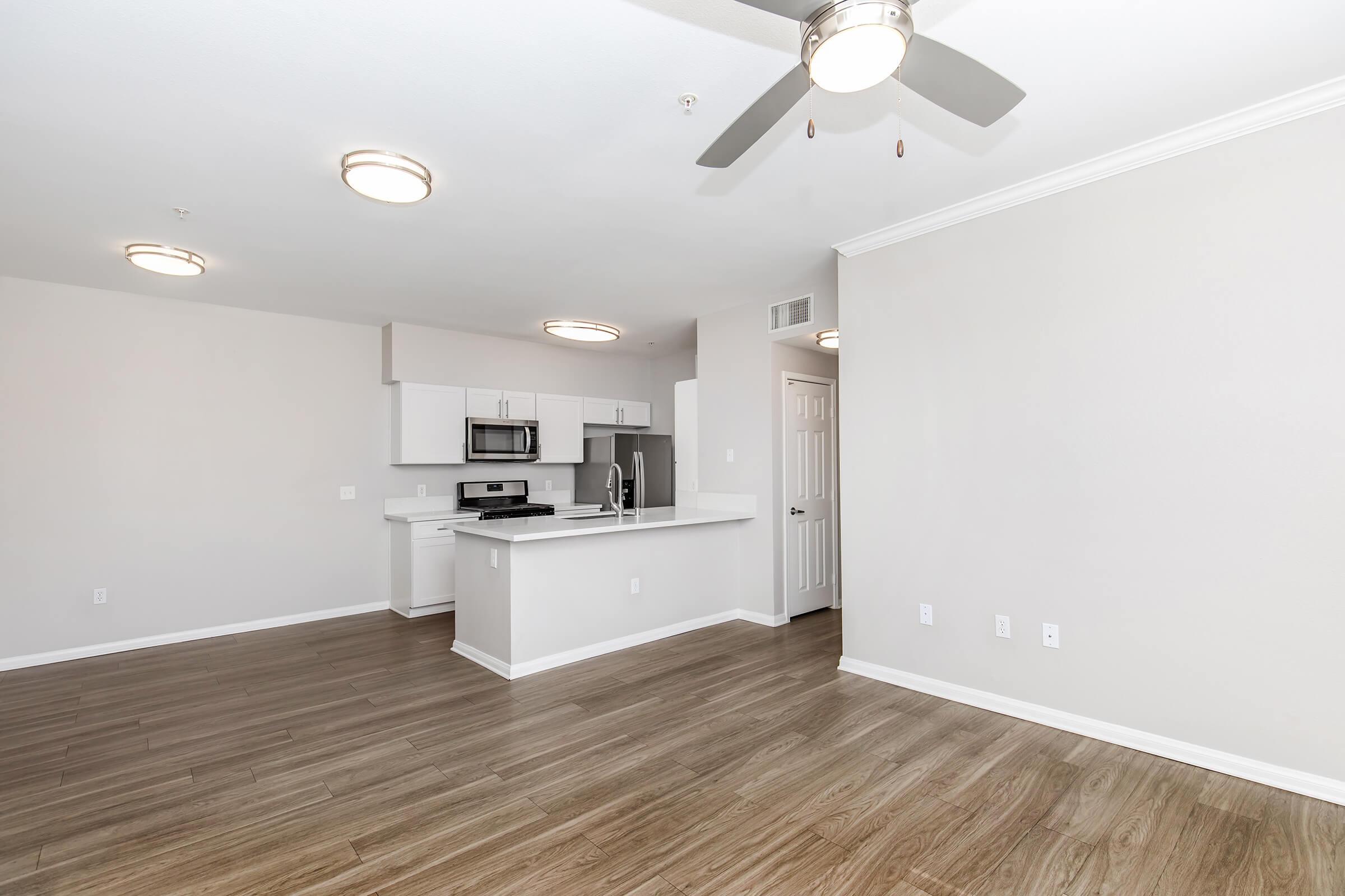 Spacious modern apartment interior featuring an open floor plan with hardwood-style flooring, ceiling fan, and ample natural light. The kitchen includes white cabinetry, stainless steel appliances, and a breakfast bar, while the living area has a neutral wall color and light fixtures.
