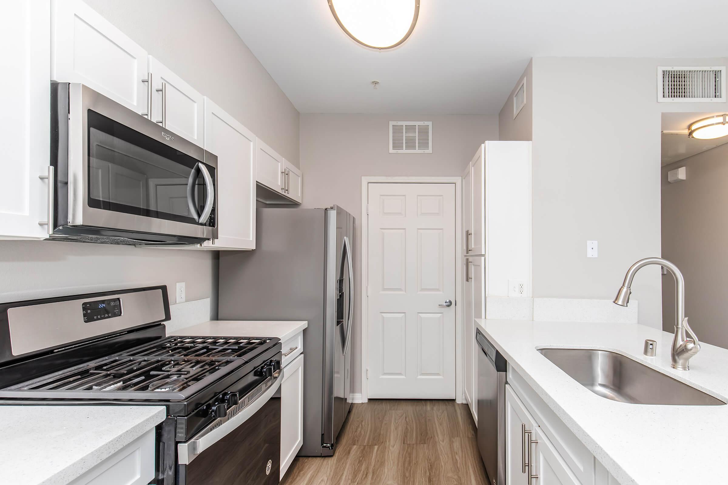 Modern kitchen featuring white cabinets, stainless steel appliances including a gas stove and microwave, a refrigerator, and a stone countertop. There’s a double sink with a faucet, and the room has neutral wall colors and wood-style flooring. A door leads to an adjacent area.
