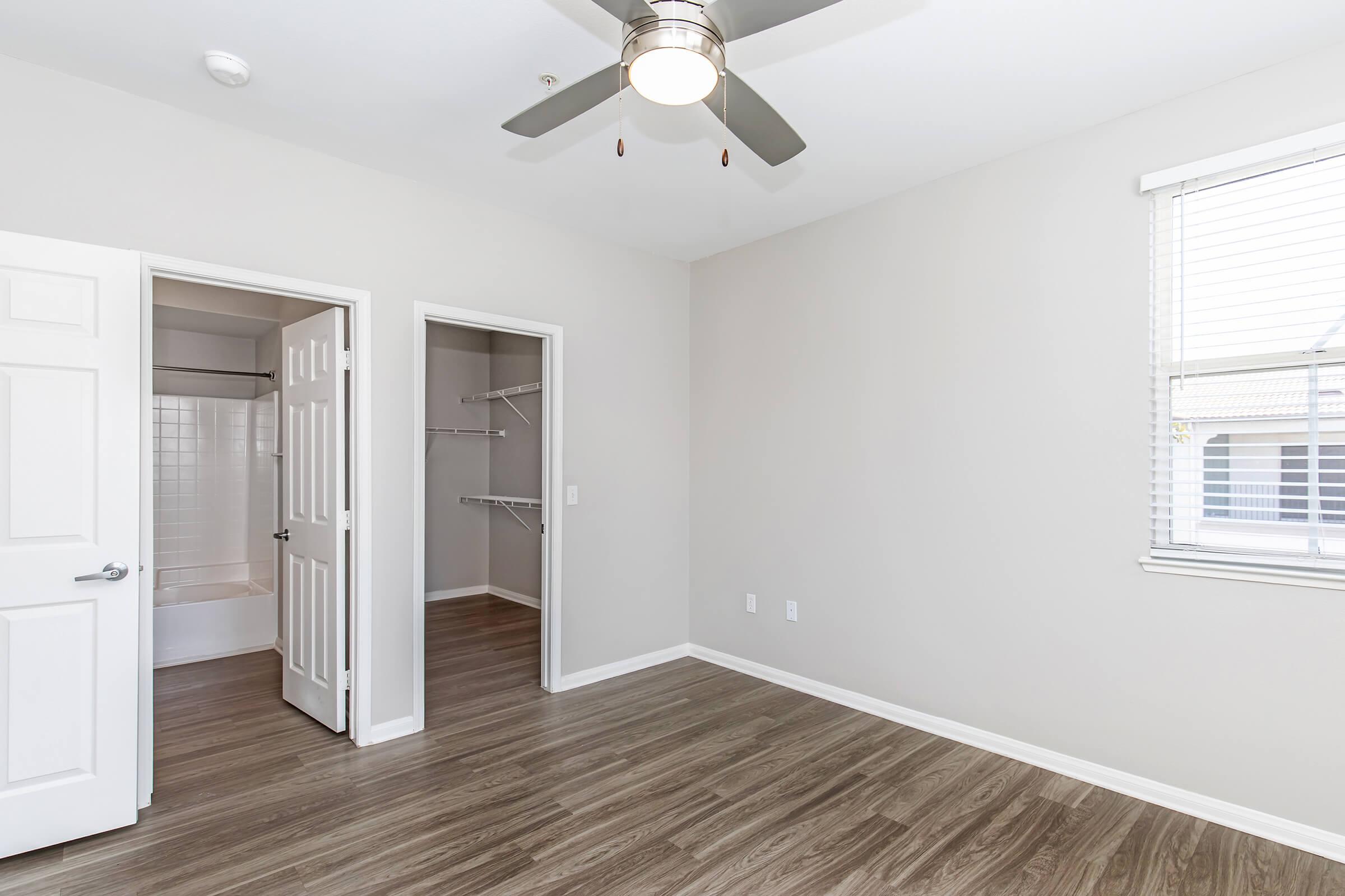 A clean, empty bedroom featuring light gray walls, a ceiling fan, and wooden flooring. One wall has a window with white blinds, while an open door leads to a bathroom with a shower. Another door opens to a closet with shelving, providing ample storage space.