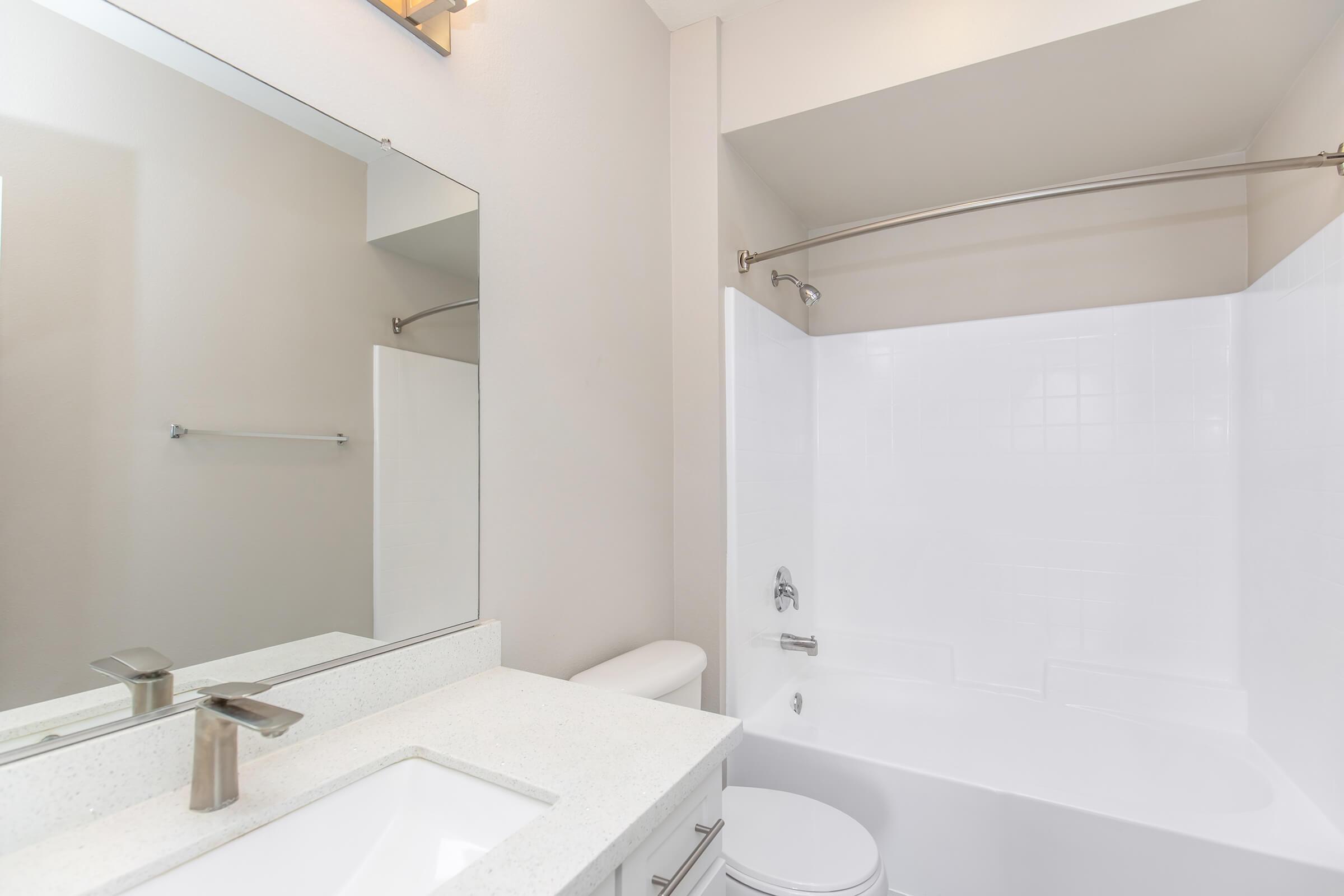 A clean and modern bathroom featuring a white bathtub with a shower, a large mirror above a sleek countertop with a sink, and a minimalistic design. The walls are painted in a light color, providing a bright and airy atmosphere. A towel bar is visible on the wall beside the shower.