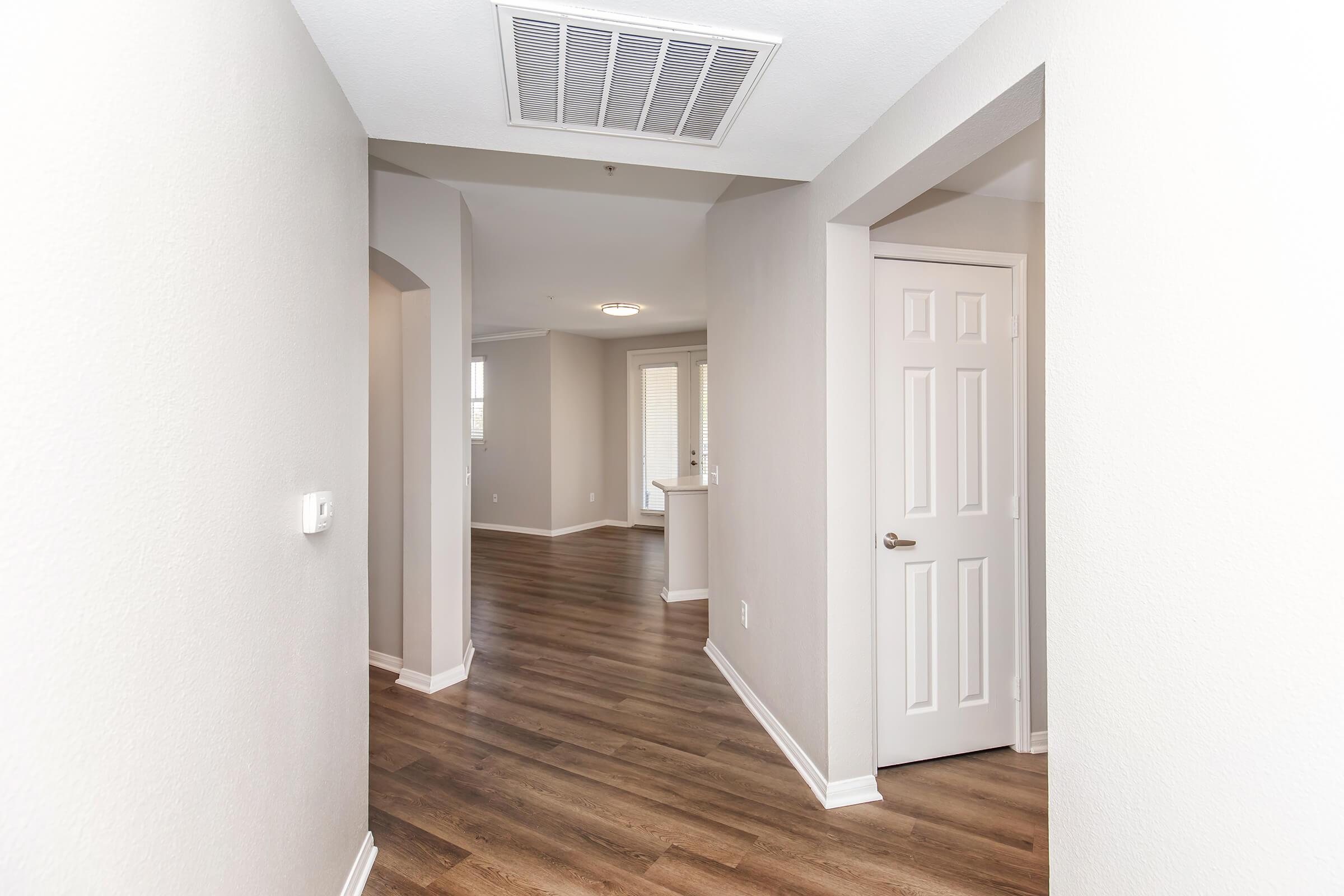 A well-lit interior hallway with light gray walls and wooden flooring. The hallway extends towards a doorway on the right leading to a room, with an archway on the left. At the end, a glimpse of a bright living space is visible. Soft ceiling lights and a vent can be seen above.