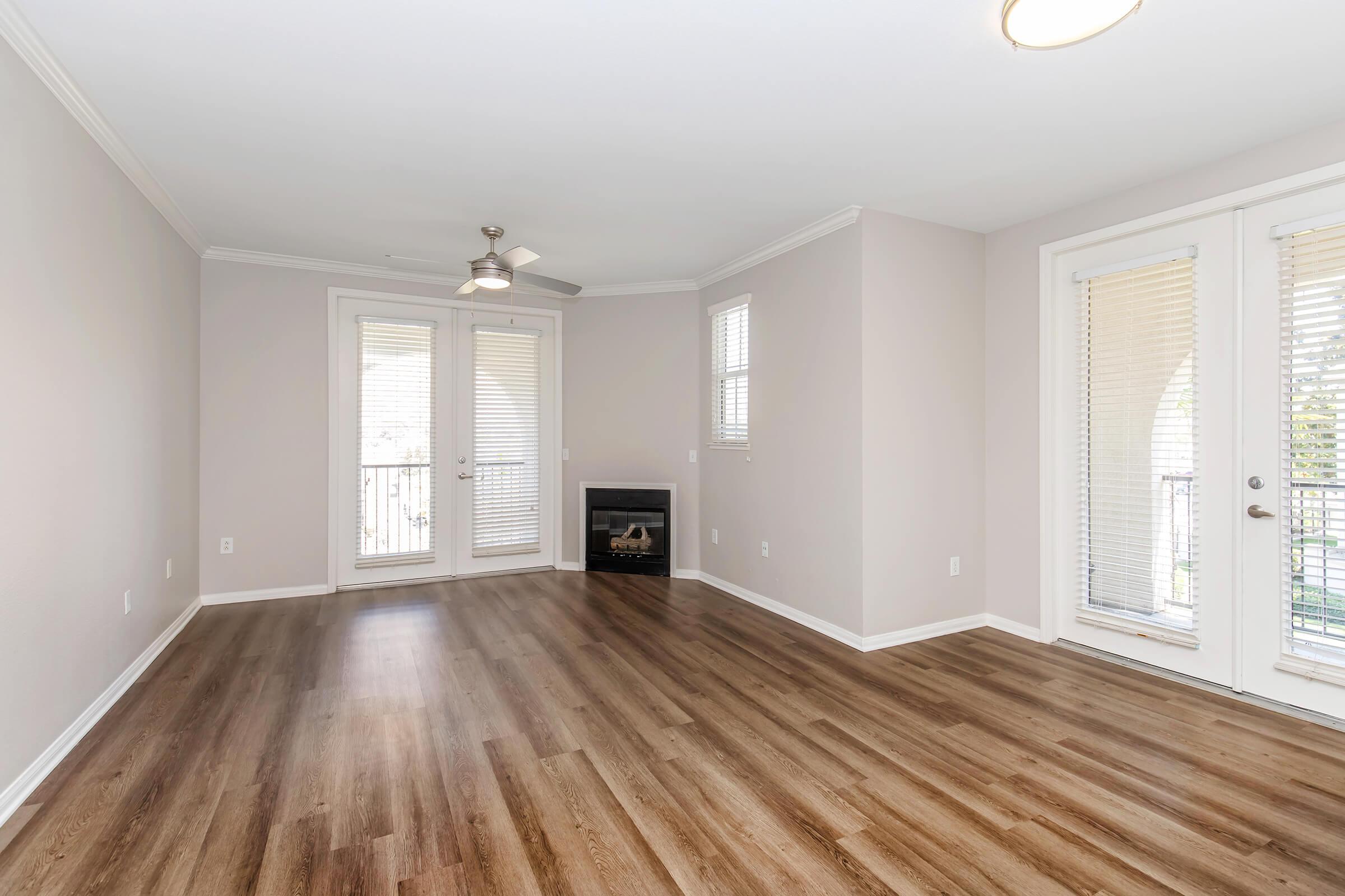 A spacious, well-lit living room featuring light beige walls, a ceiling fan, and dark wood laminate flooring. There are large windows with white blinds and two glass doors leading to an outdoor balcony. A small electric fireplace is located in one corner, enhancing the cozy atmosphere.
