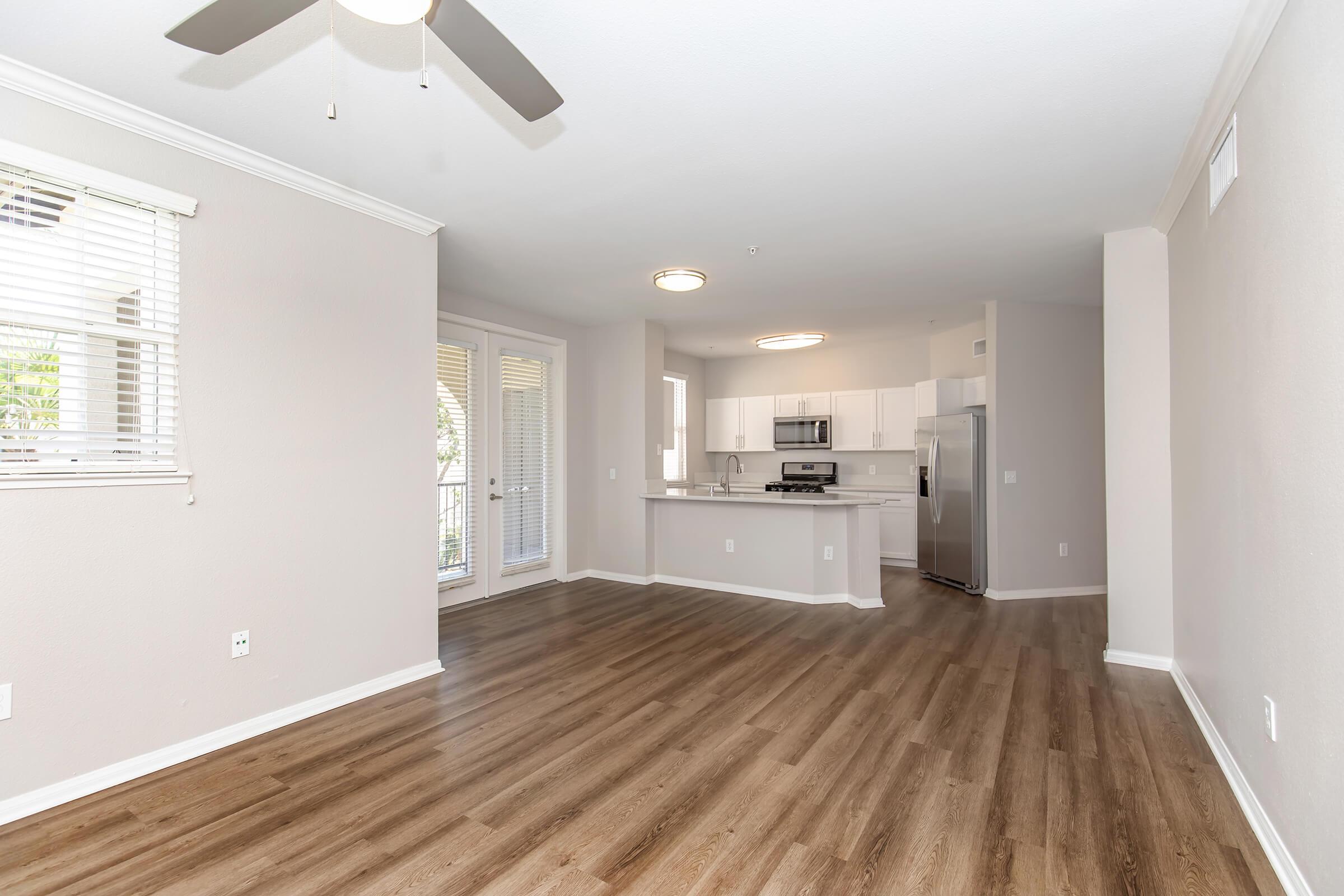 Spacious modern living area featuring light-colored walls, wood-like flooring, and large windows. A ceiling fan is installed, and the open layout connects to a kitchen with stainless steel appliances and a breakfast bar. Natural light brightens the room, creating a welcoming atmosphere.