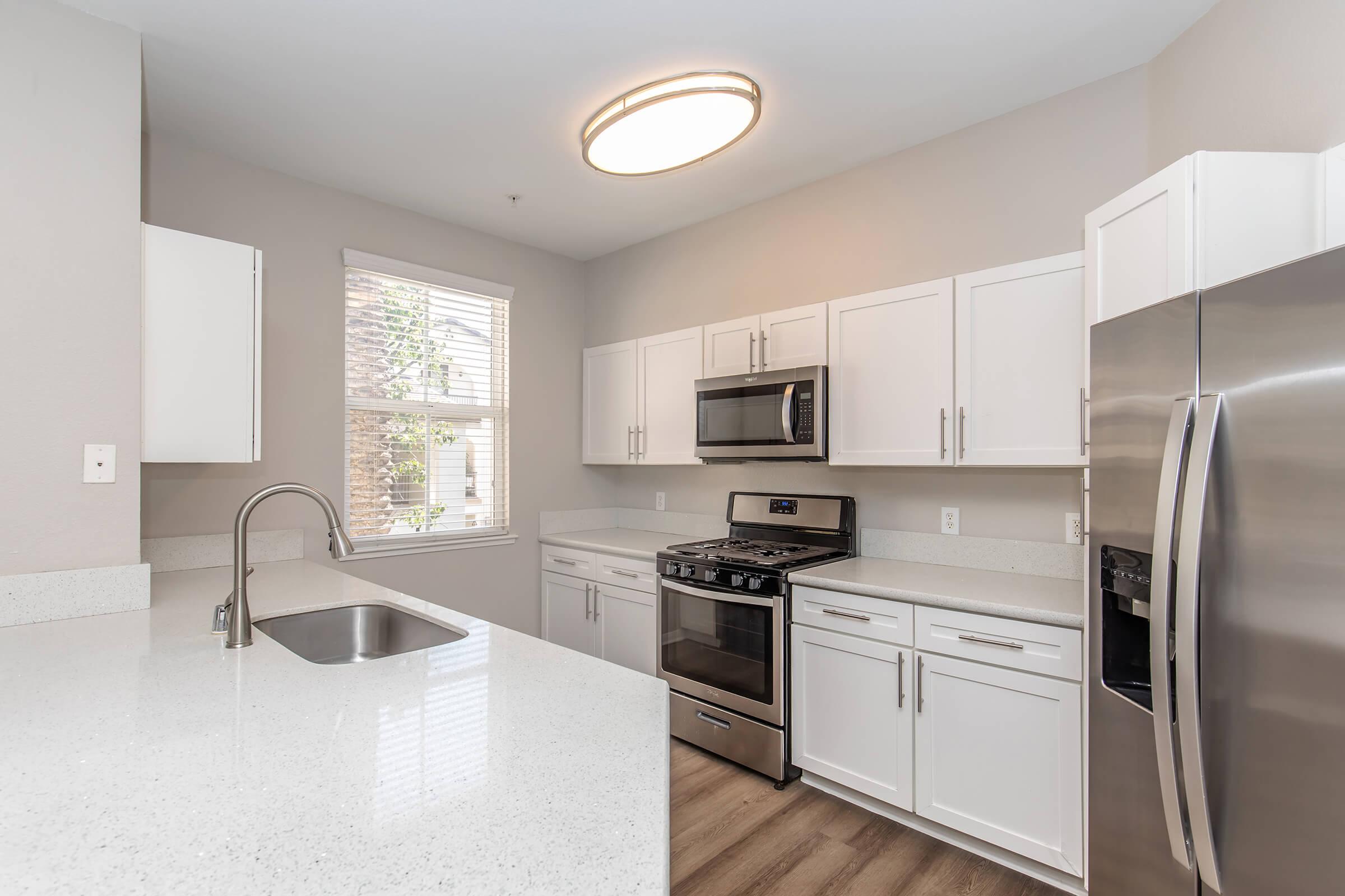 Modern kitchen featuring white cabinets, stainless steel appliances including a refrigerator and gas stove, and a double sink. The room has a neutral-colored wall and a bright overhead light, with a window allowing natural light to enter. The floor is covered with light wooden laminate.