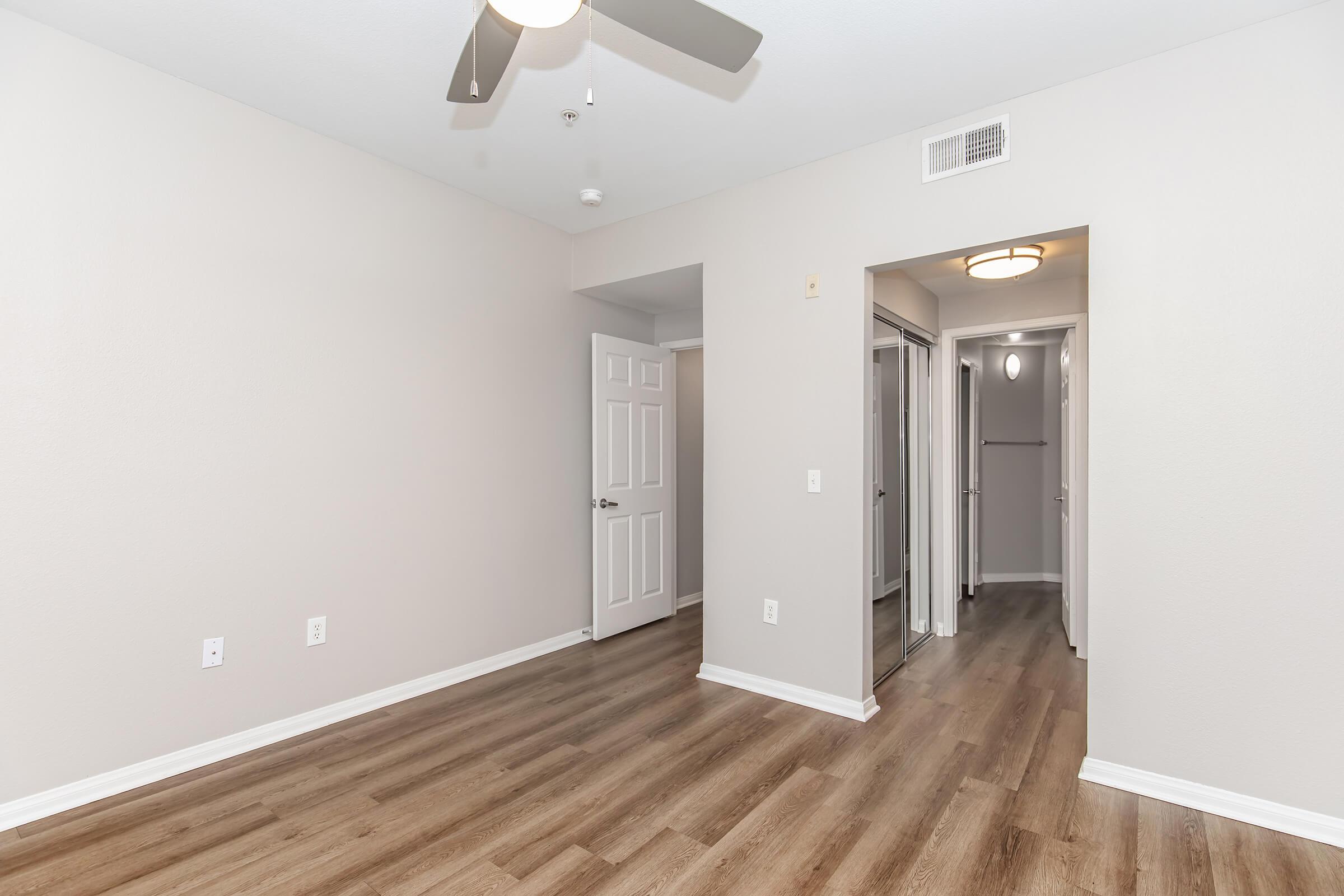 A spacious room with light-colored walls and wooden flooring. There is a ceiling fan, a closet on the right, and an open doorway leading to another area. The overall design is modern and clean, with ample natural light.