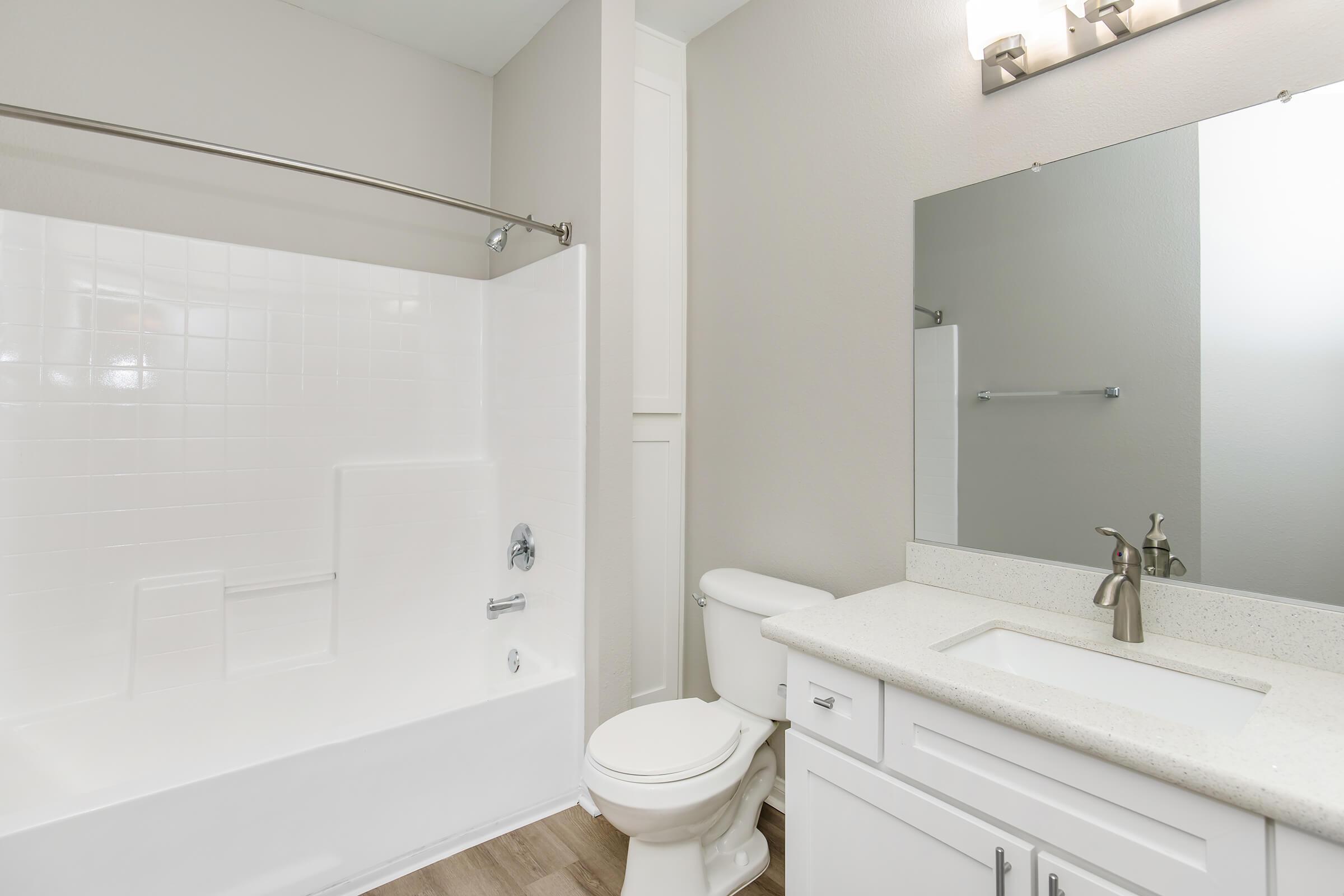 A modern bathroom featuring a white bathtub and shower combination, a toilet, and a vanity with a sink. The walls are painted in a light color, and there is a large mirror above the sink. Soft lighting is provided by two sconces on the wall. The floor is a laminate wood style.