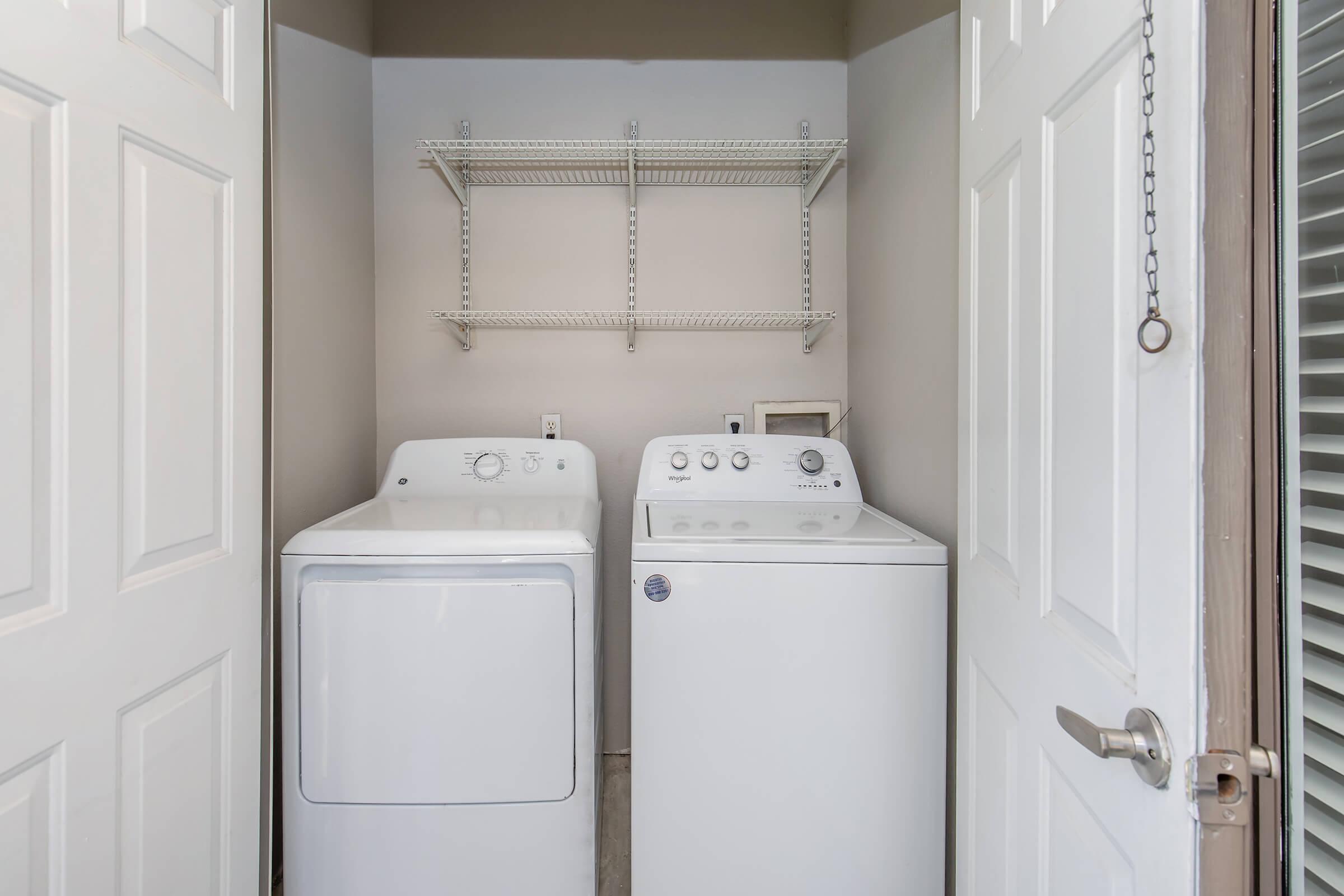 A laundry room featuring a white washing machine and a white dryer side by side. Above them is a wire shelf for storage. The room is enclosed with double doors, giving a tidy appearance with neutral-colored walls.