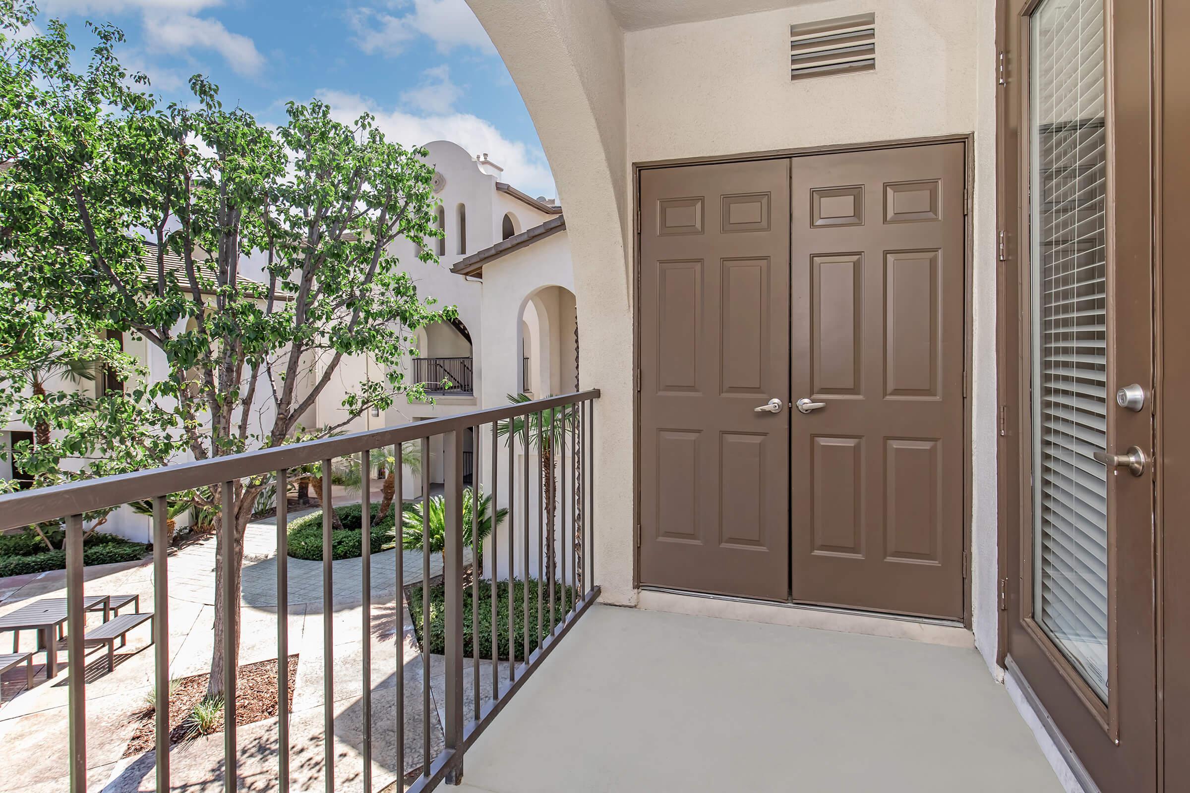 A view of a balcony with a railing overlooking a landscaped courtyard. The scene includes two closed brown doors leading to an apartment, a tree with green leaves nearby, and a bright sky with clouds in the background. The architecture features arched doorways and a light-colored exterior.