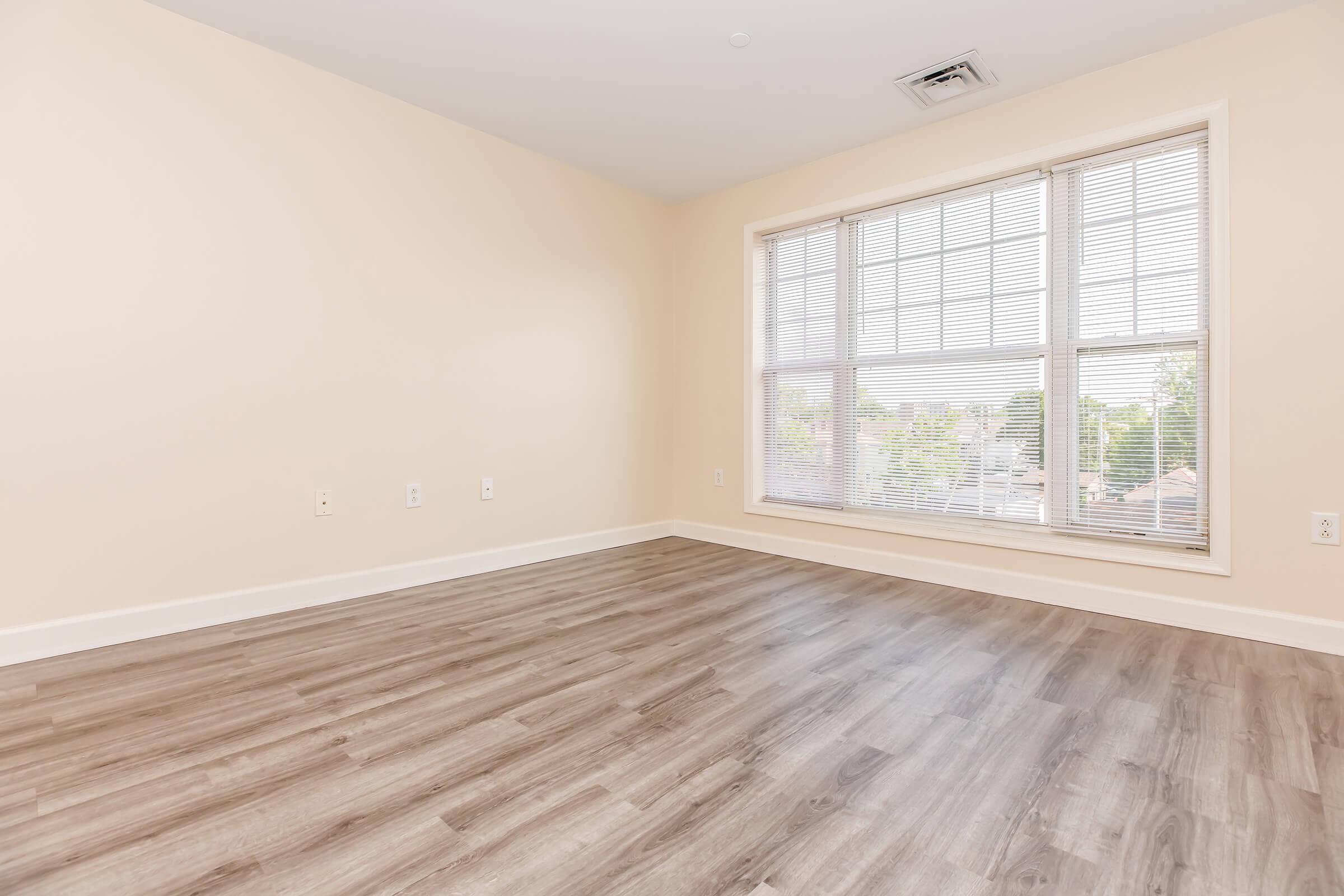 Empty room with neutral beige walls and light-colored wooden flooring. A large window with white blinds allows natural light to fill the space, highlighting the simplicity of the room. The area is uncluttered, creating a blank canvas for potential decoration or furnishing.