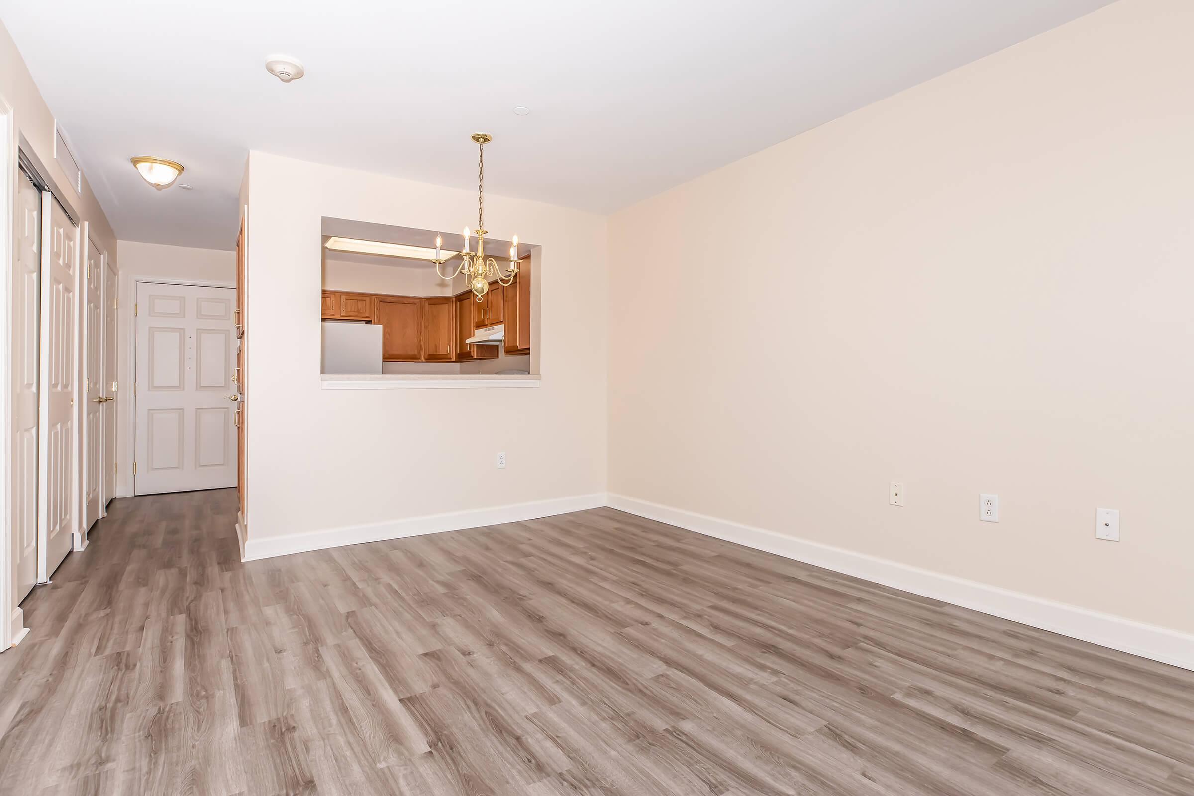 Spacious, empty room with light beige walls and laminate flooring, featuring a small chandelier and an open view into a kitchen area through an archway. Doors lead to additional rooms on the left side, with natural light illuminating the space.