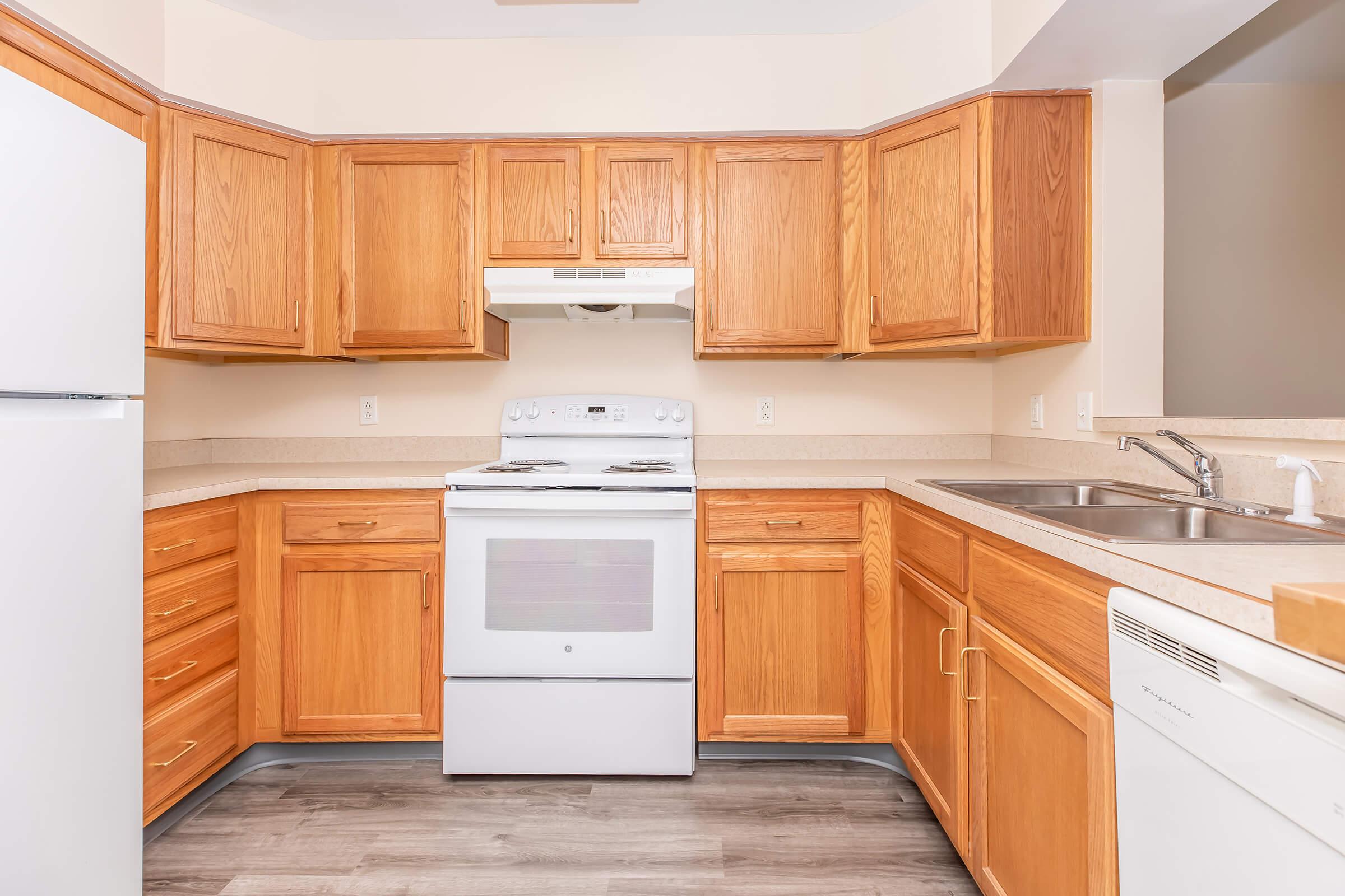 A modern kitchen featuring wooden cabinetry, a white stove and hood, a double sink, and a refrigerator. The countertops are light-colored, and the flooring is a gray laminate. The space is well-lit and organized, with an open design that connects to adjacent areas.