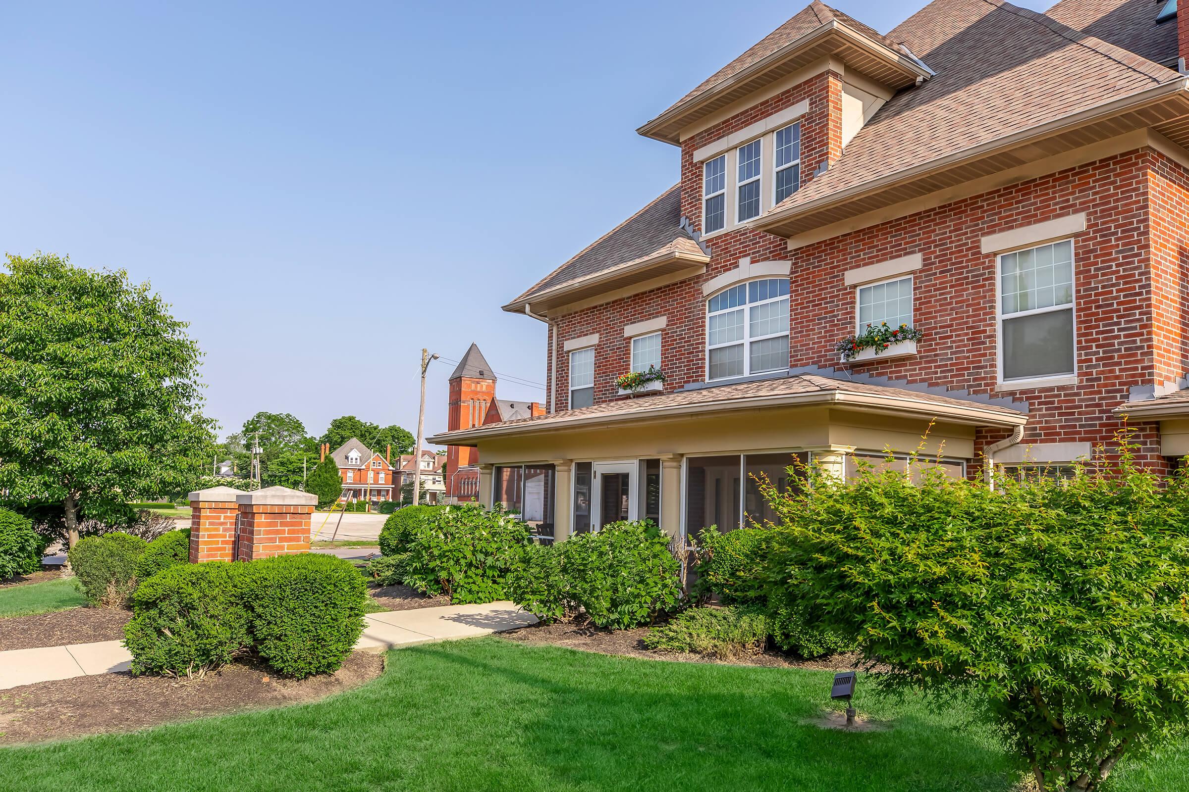 A well-maintained brick residential building with multiple windows and flower boxes, surrounded by lush green landscaping. A walkway leads to the entrance, and in the background, another building can be seen along with a distant tower. The sky is clear and blue, indicating a bright day.