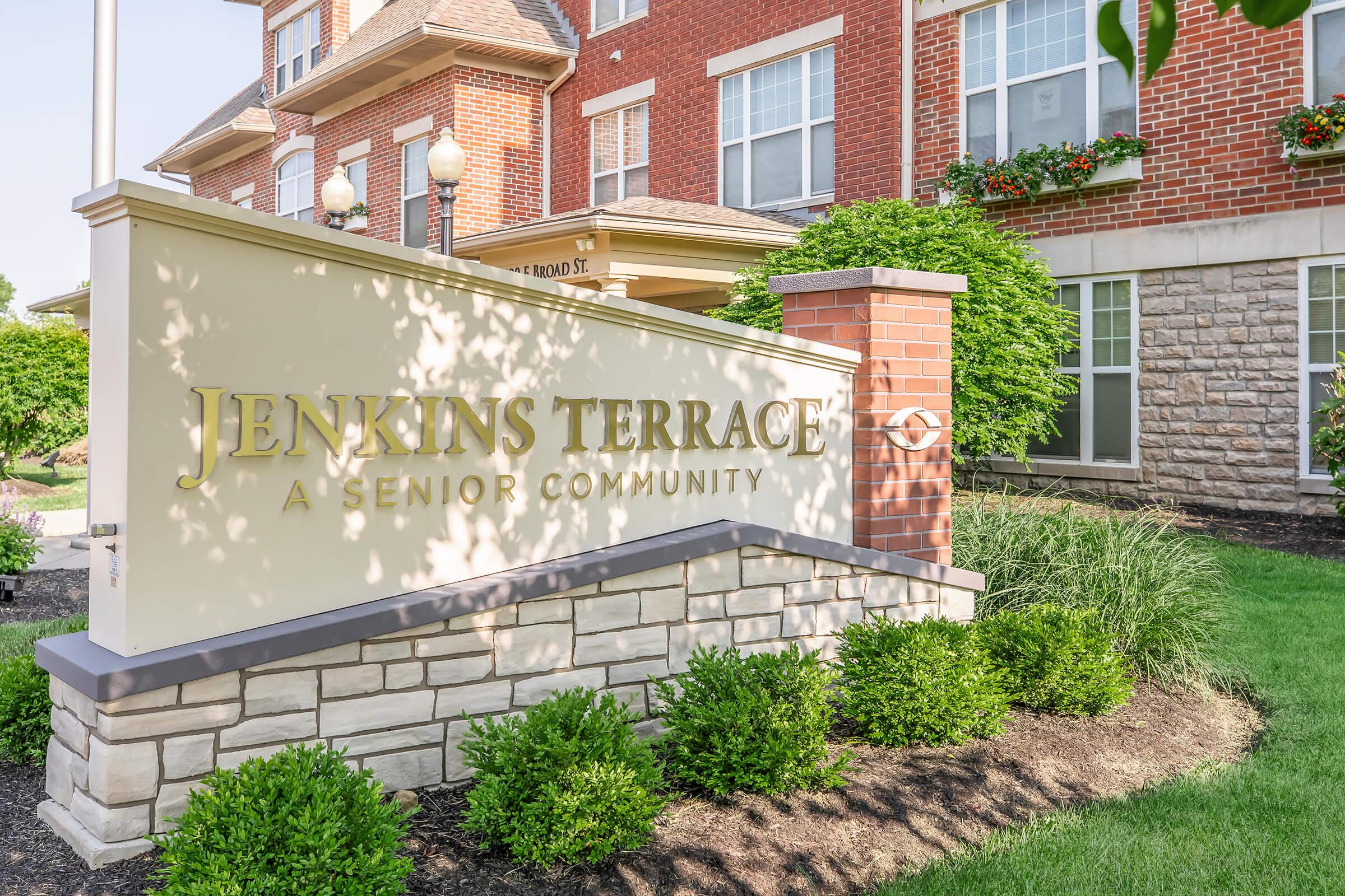 Sign featuring the text "Jenkins Terrace, A Senior Community," with landscaping elements such as bushes and a decorative brick structure. The background shows a well-maintained building, creating a welcoming atmosphere for residents and visitors.