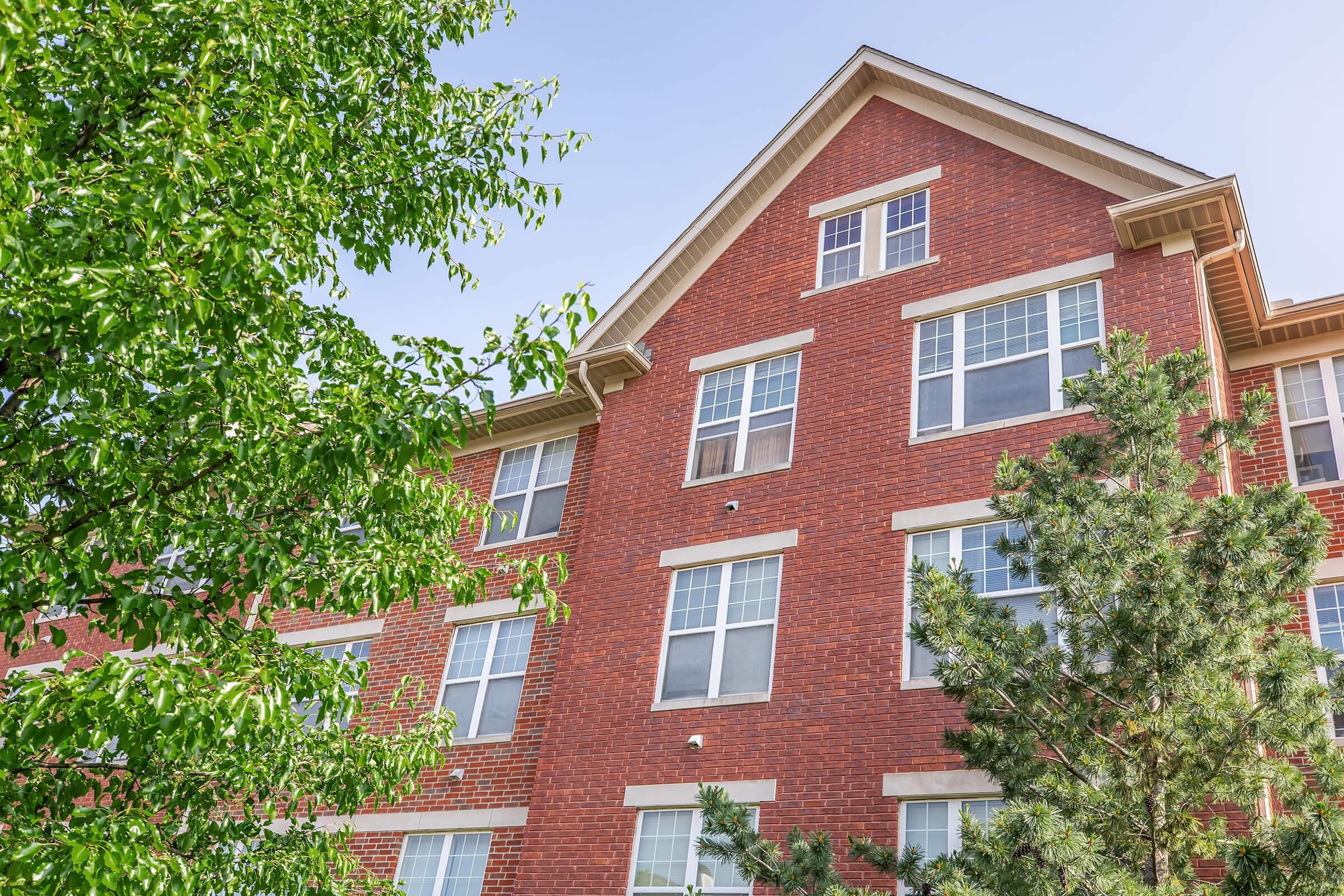 A close-up view of a multi-story brick apartment building with several windows and a tree in the foreground, showcasing a clear blue sky. The structure features a peaked roof and is surrounded by greenery, providing a welcoming residential atmosphere.