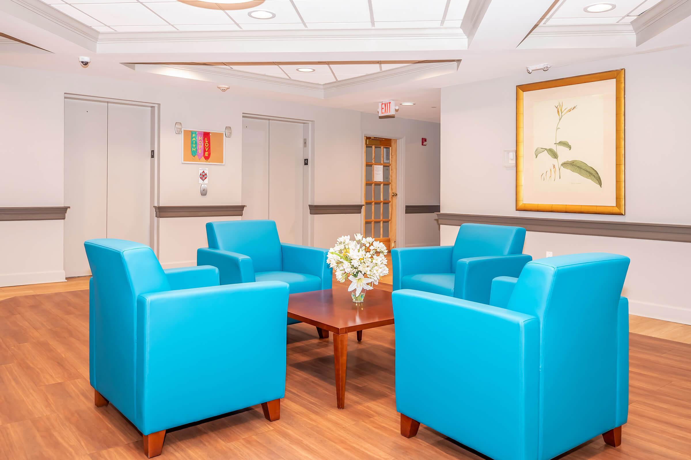 Brightly lit waiting area featuring four turquoise chairs arranged around a wooden table with a vase of white flowers. The walls are light-colored, and there are elevators to the left, along with a framed botanical artwork on the right. The space has a clean, modern look.