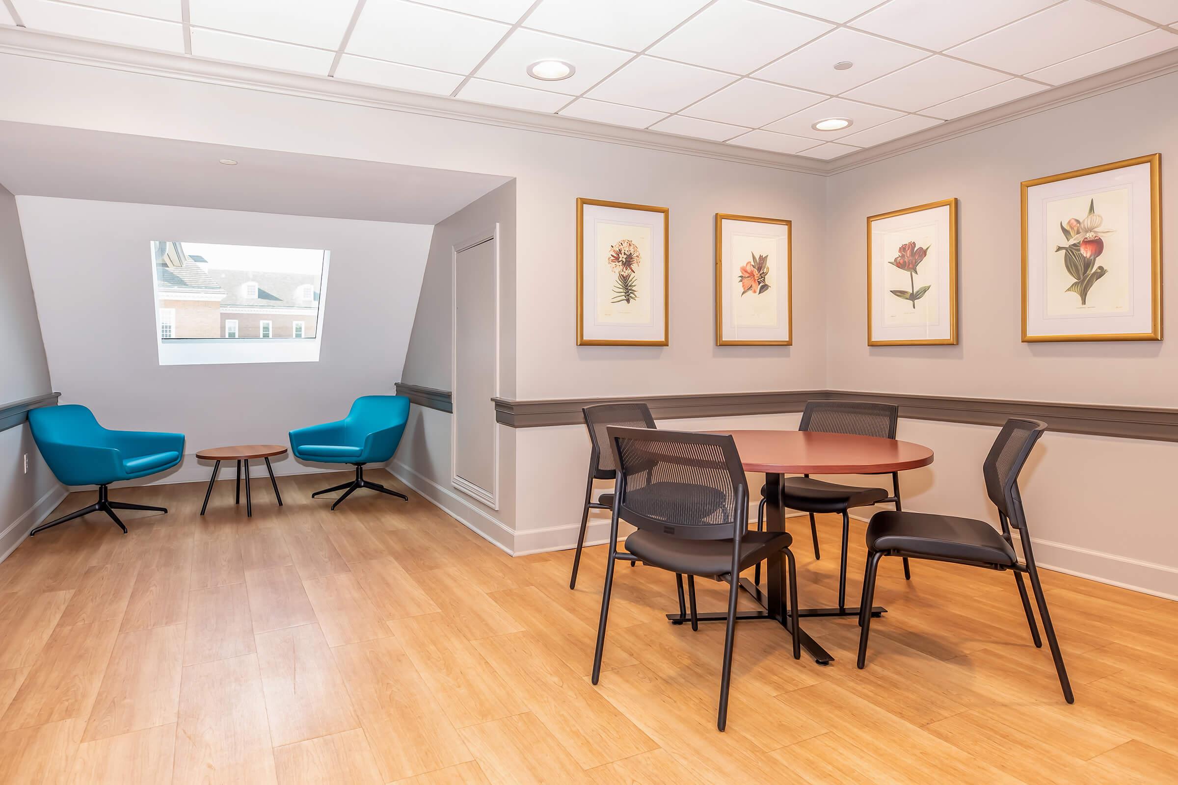A cozy corner of a bright room featuring a round table surrounded by black chairs. Two stylish blue armchairs are positioned near the window, and framed floral artwork decorates the walls, contributing to a tranquil atmosphere. The wooden floor adds warmth to the modern design.