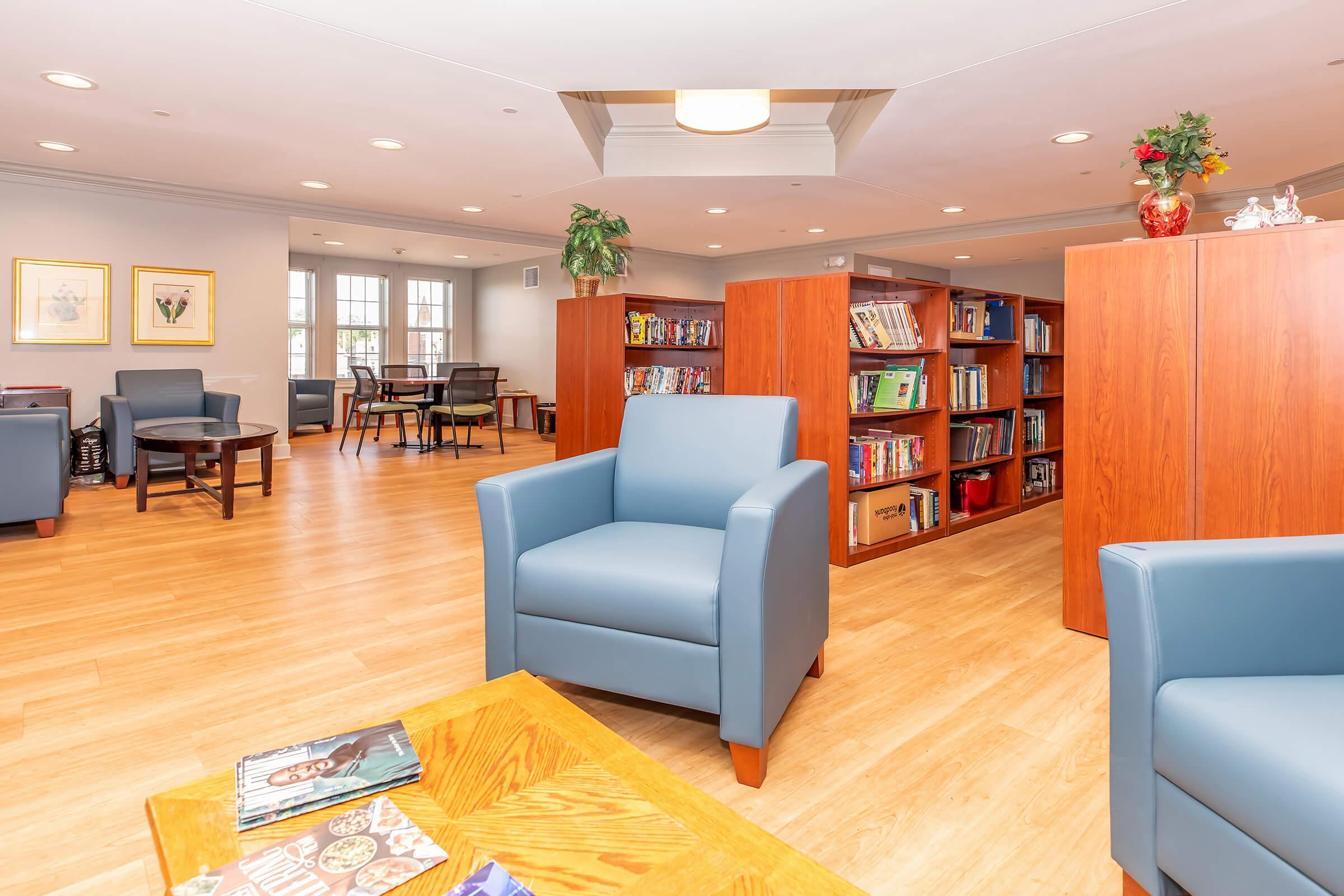 A cozy indoor library or reading area featuring wooden bookshelves filled with books, comfortable blue chairs, and a coffee table. Natural light flows in through large windows, creating a welcoming atmosphere for relaxation and reading.