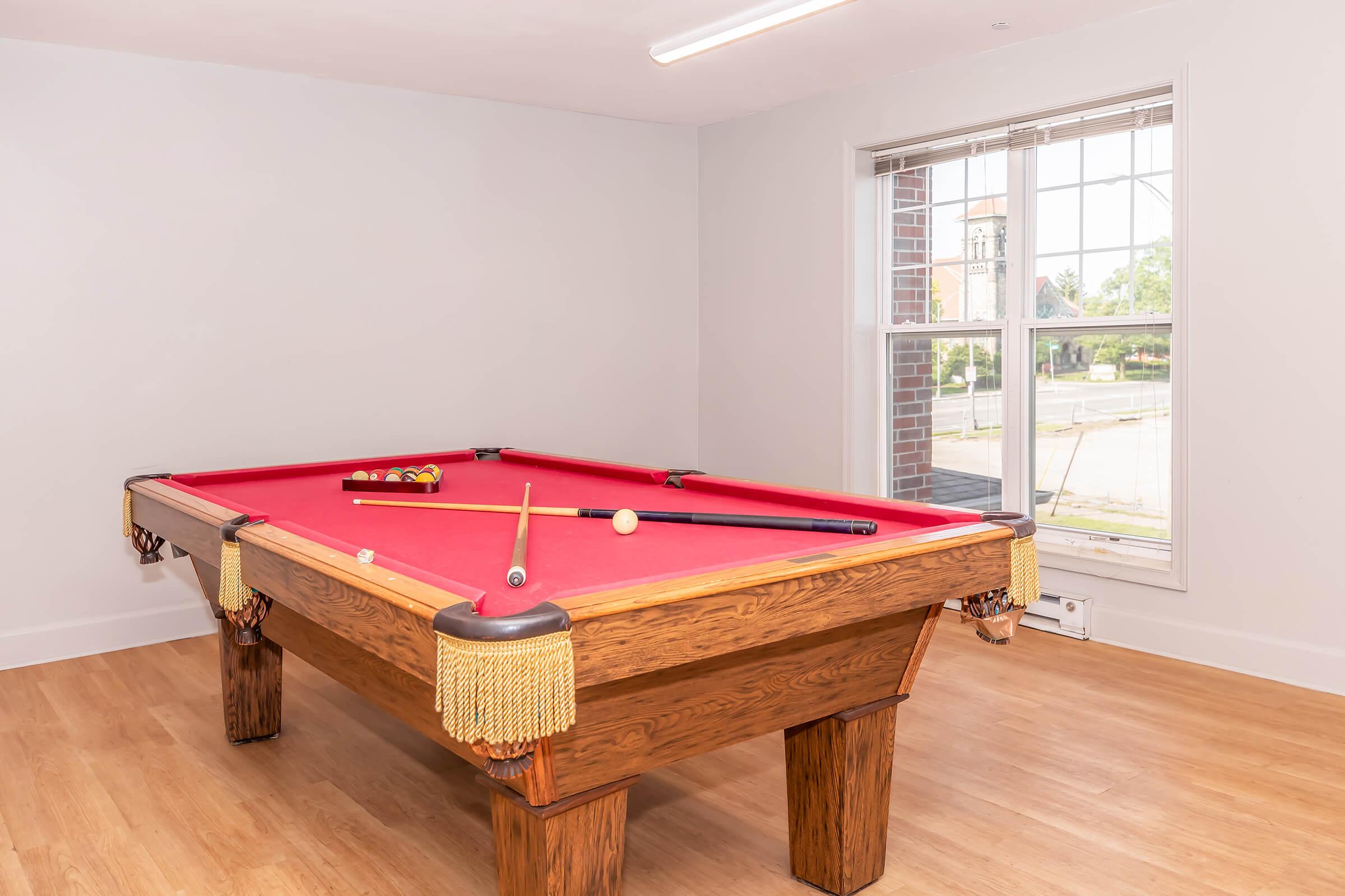 A red felt billiards table with wooden legs, featuring a cue ball, a cue stick, and several colored pool balls scattered on the table. The room has light wood flooring and a large window with a view of a building outside, providing natural light.