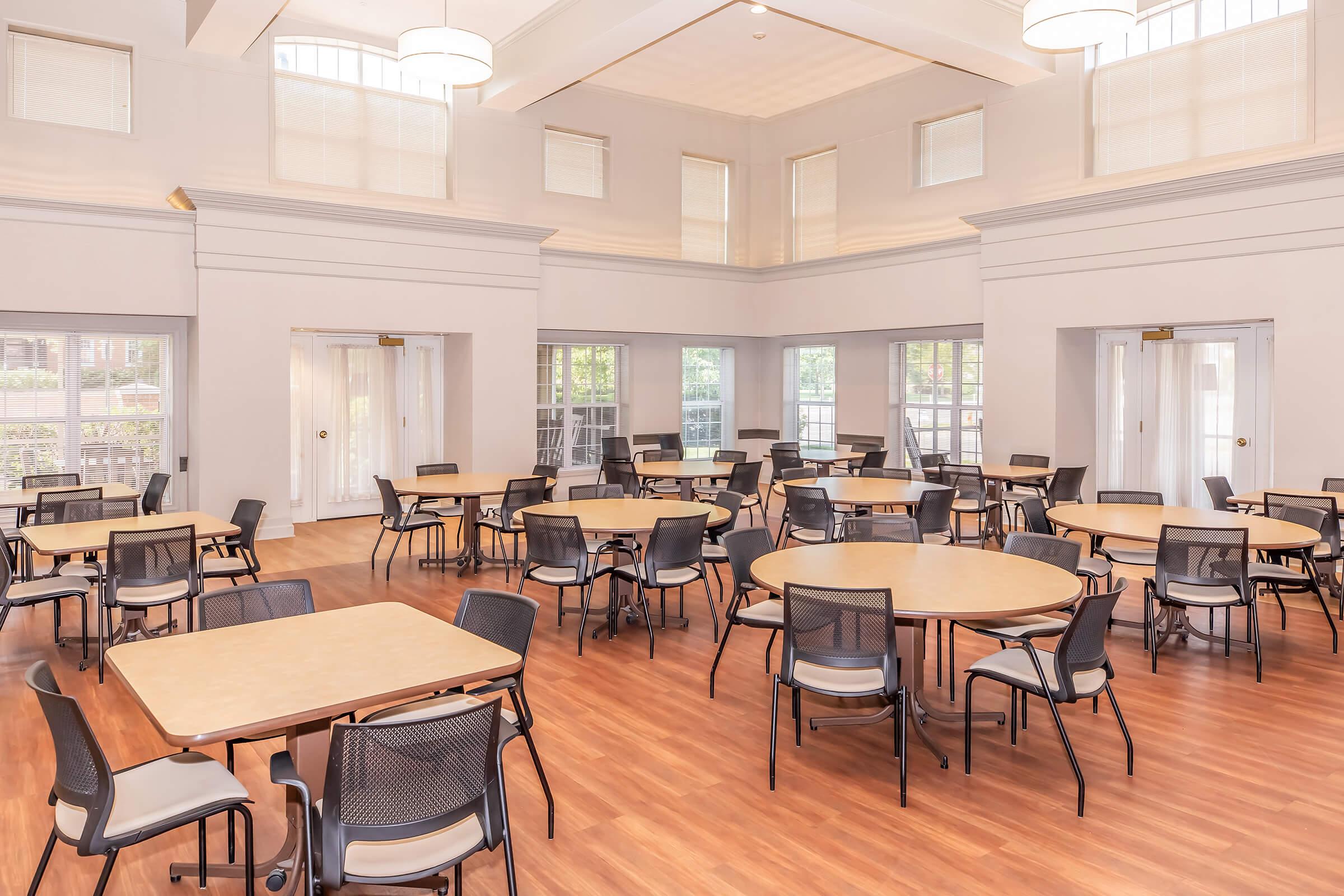 Empty dining hall with multiple round tables and chairs arranged neatly. Large windows allow natural light to fill the space, highlighting the wooden floor. The room features a high ceiling with decorative light fixtures, creating an inviting atmosphere suitable for gatherings or events.