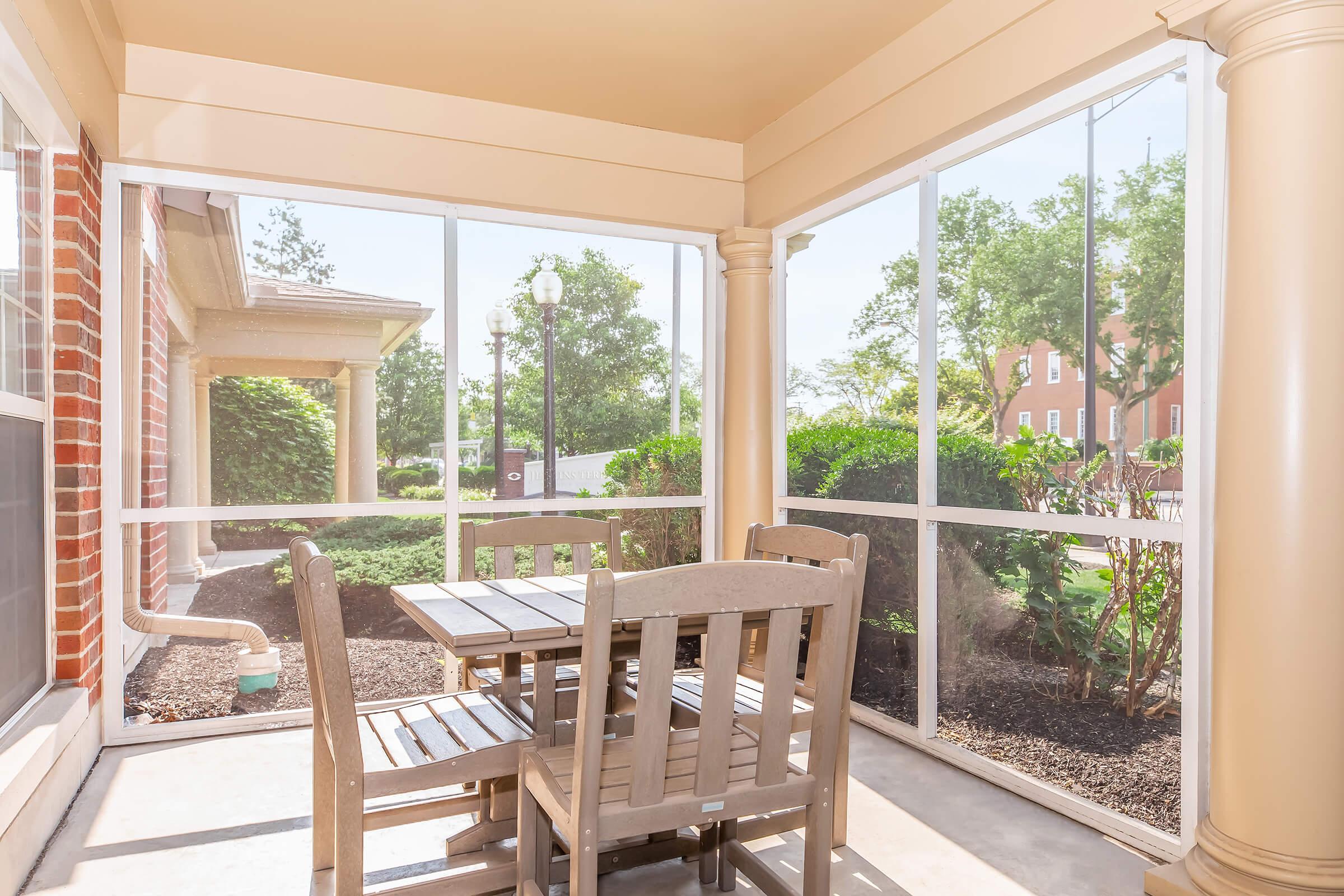 A screened-in porch featuring a wooden table and chairs, with a view of a garden and landscaped surroundings. The space is well-lit with natural light filtering through the screens, and columns accentuate the entrance.