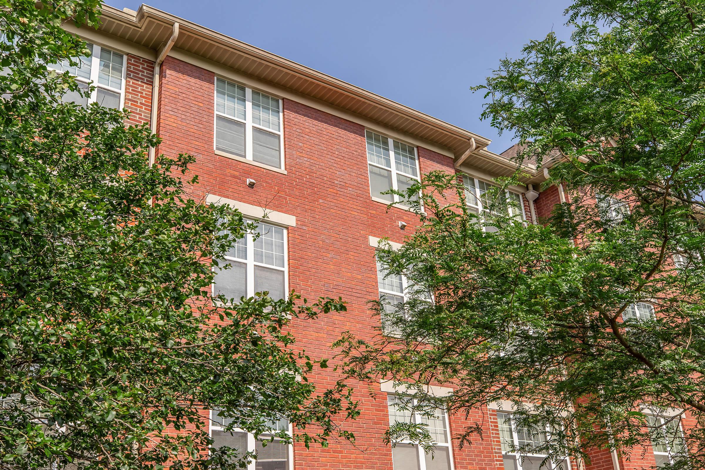 A view of a red-brick apartment building with multiple windows, framed by green trees. The building features several floors and a clear blue sky in the background, creating a picturesque residential setting.