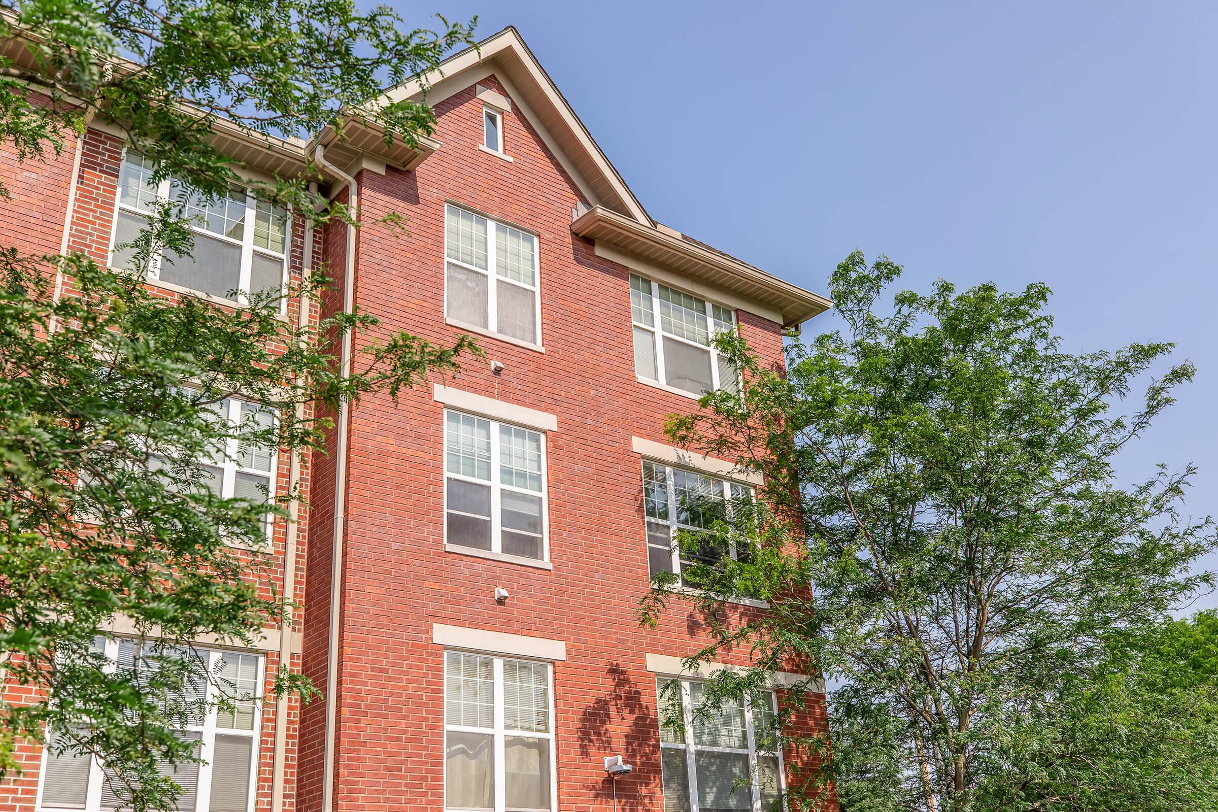 A red brick apartment building with multiple windows and greenery in front. The structure has three visible floors, showcasing a clear blue sky above. Trees flank the building, adding to the scene's natural ambiance.