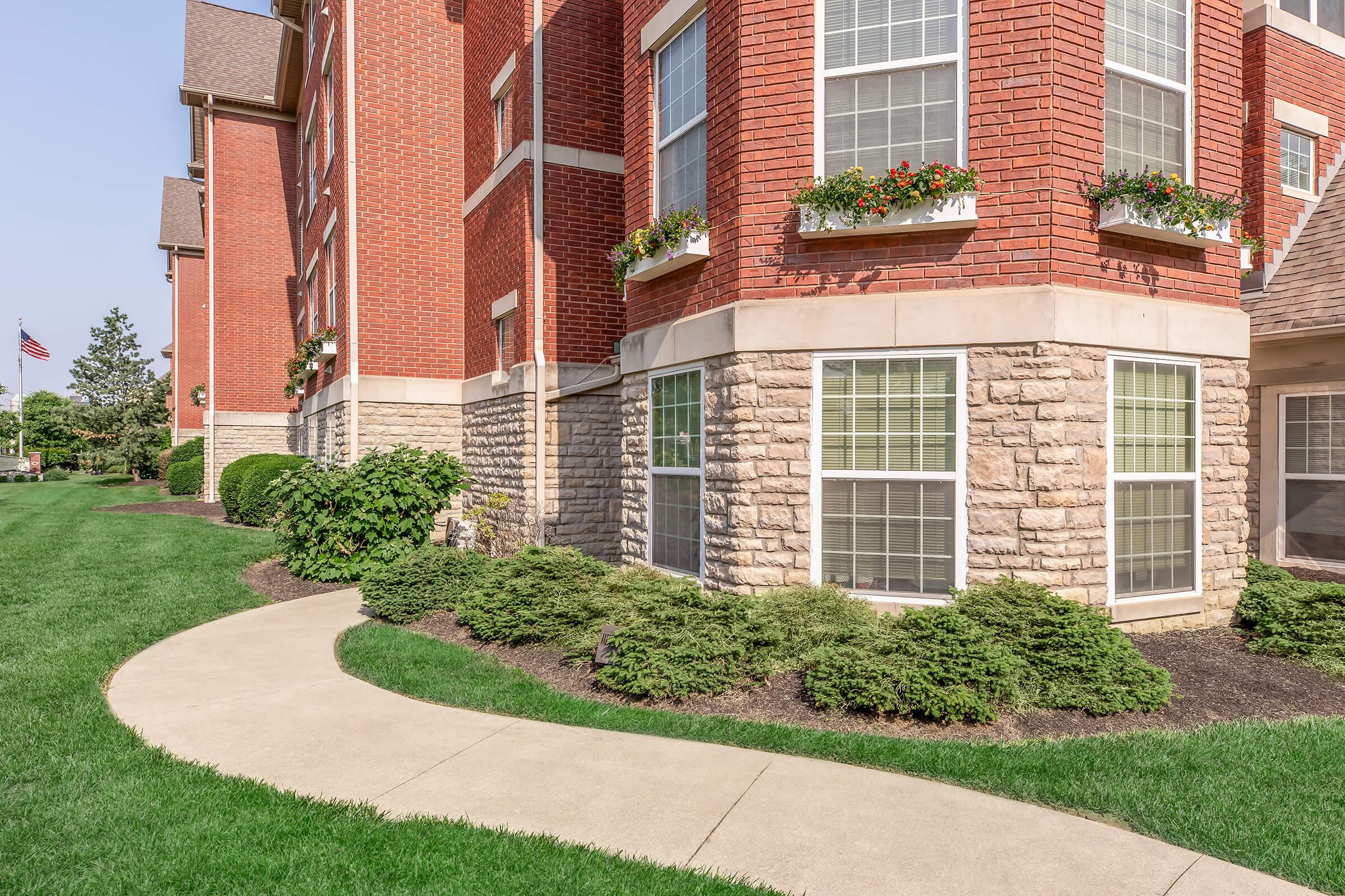 A well-maintained exterior of a brick apartment building featuring large windows adorned with flower boxes. Lush green landscaping surrounds the building, with a winding concrete pathway leading to the entrance. The scene is set under a clear blue sky.